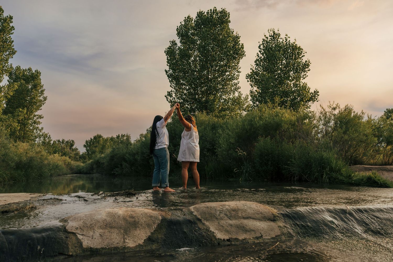 Two women dancing on rocks by a river at sunset, surrounded by lush green trees and a colorful sky.