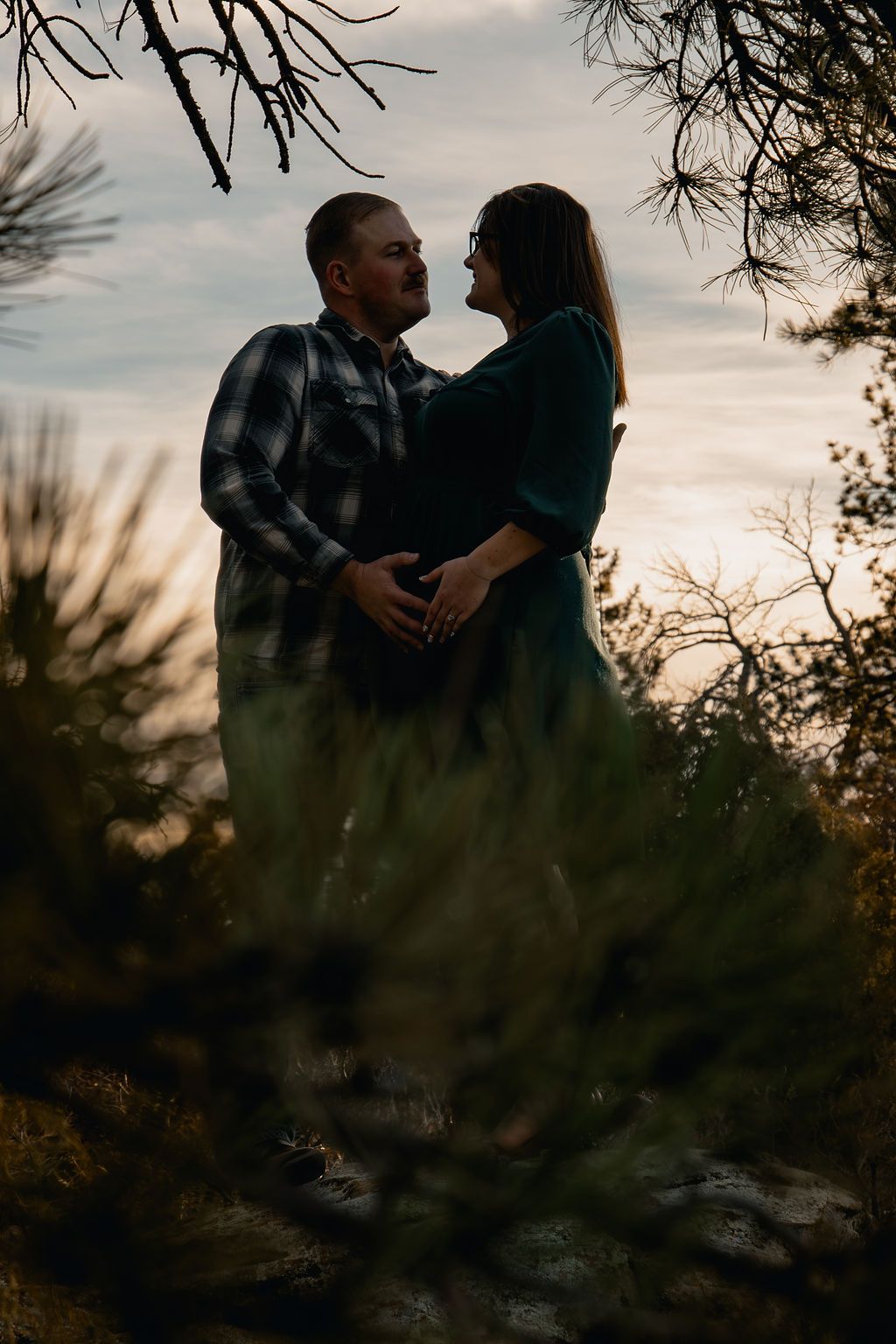 A couple embraces in silhouette against a moody sunset sky with pine tree branches framing the romantic scene.