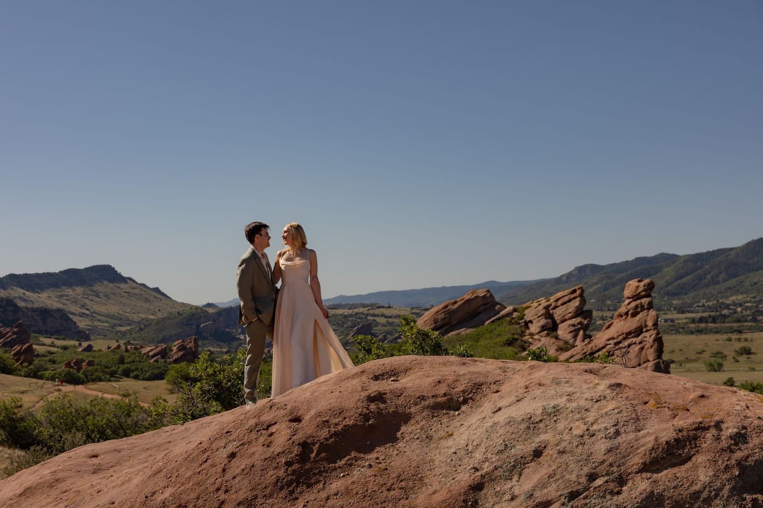 Couple in wedding attire standing on red rocks with Colorado mountain landscape and clear blue sky backdrop.