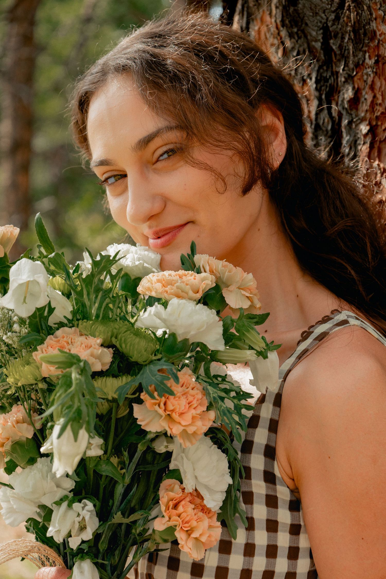 Person in gingham dress smiles while holding beautiful bouquet of peach and white carnations against tree trunk outdoors.
