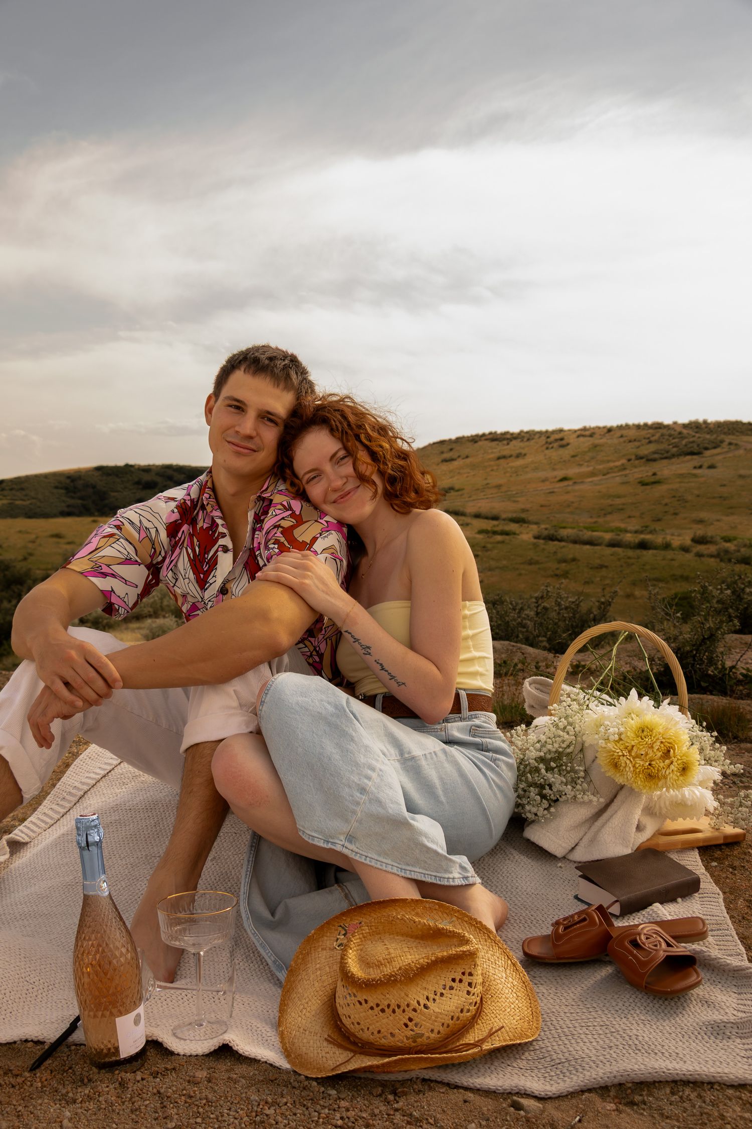 A romantic picnic setting in the countryside with a couple lounging on a blanket next to a straw hat and wildflowers.