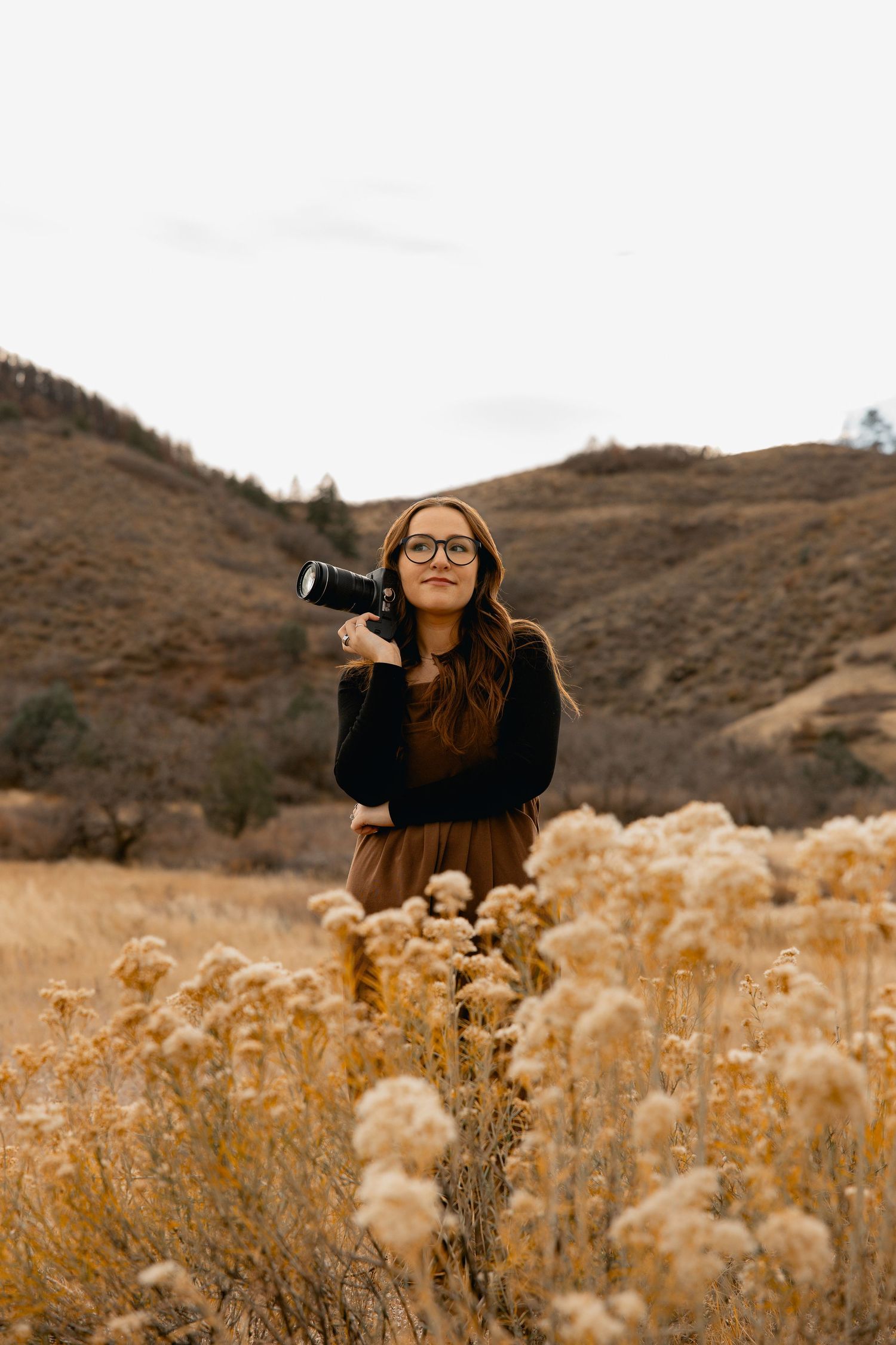 A photographer in dark clothing takes photos in a field of golden dried wildflowers with brown hills in the background.