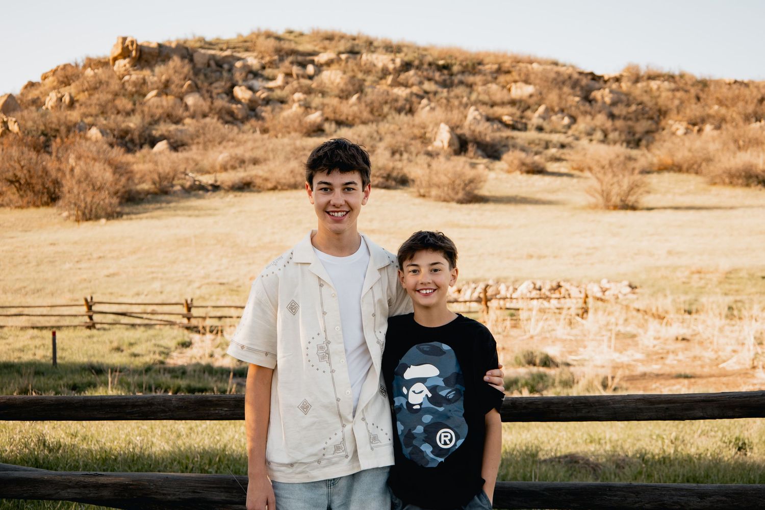 Two people stand together smiling in front of a golden hillside landscape with fence in the background.