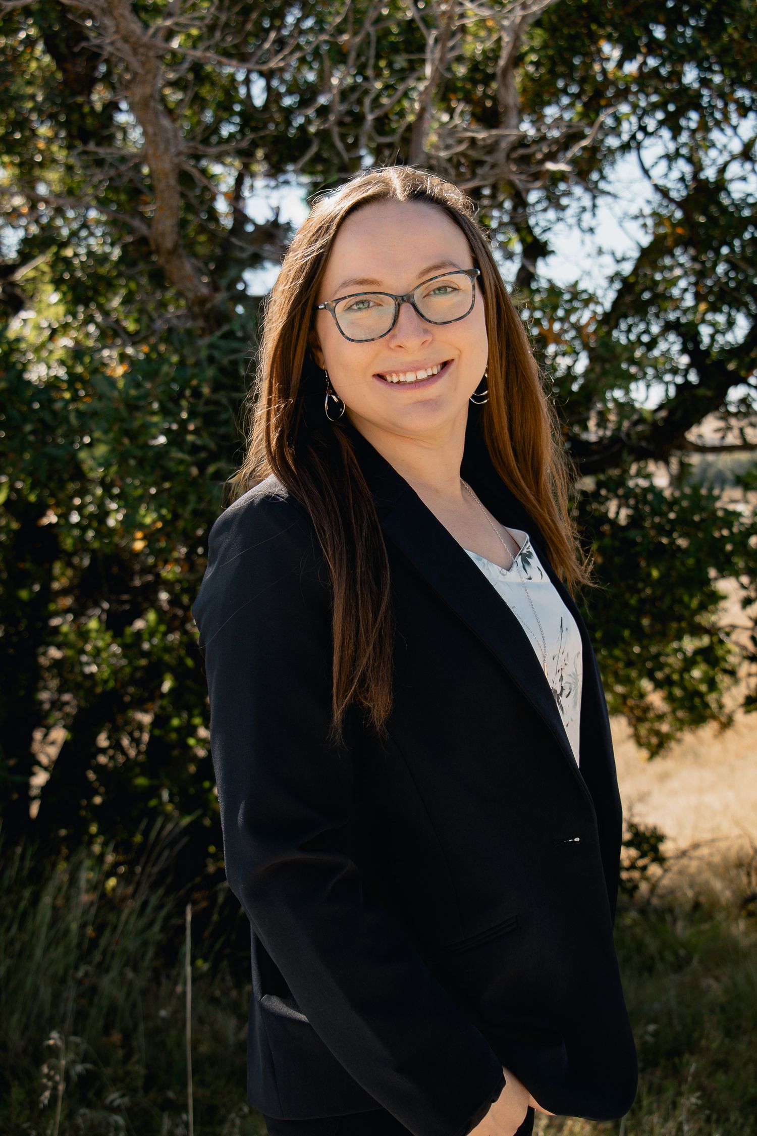 Professional outdoor portrait of a person wearing glasses and a black blazer smiling in front of trees and natural scenery.
