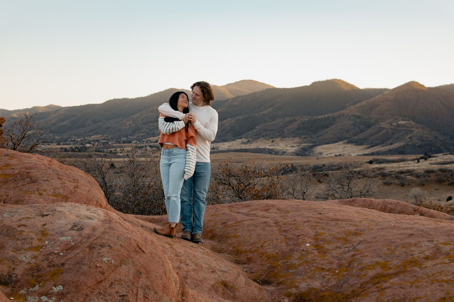 A couple embraces on red rock formations during sunset in a desert landscape with mountains in the background.