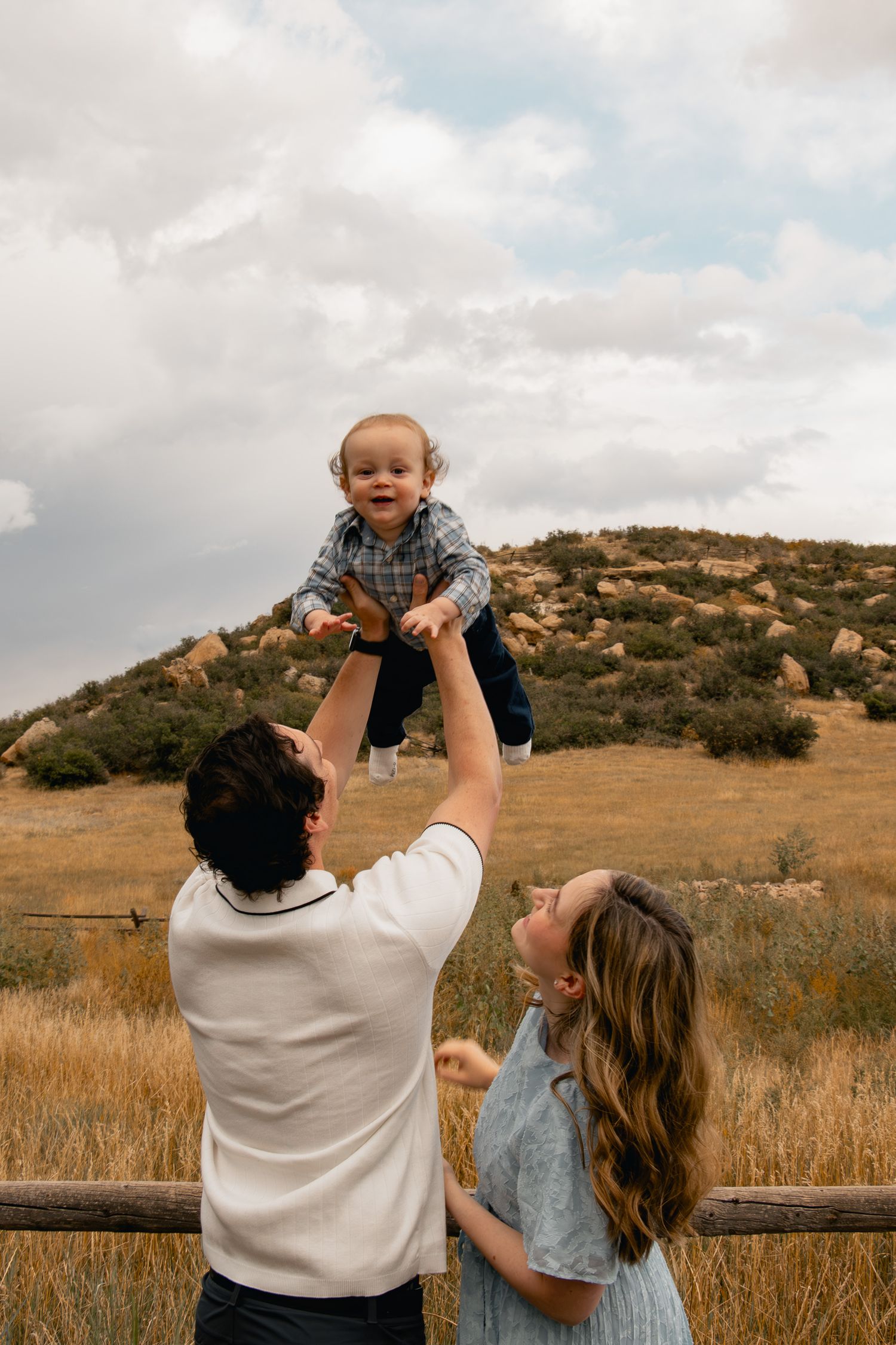 A joyful family moment captured in a golden field as the sun sets behind rolling hills in the distance.