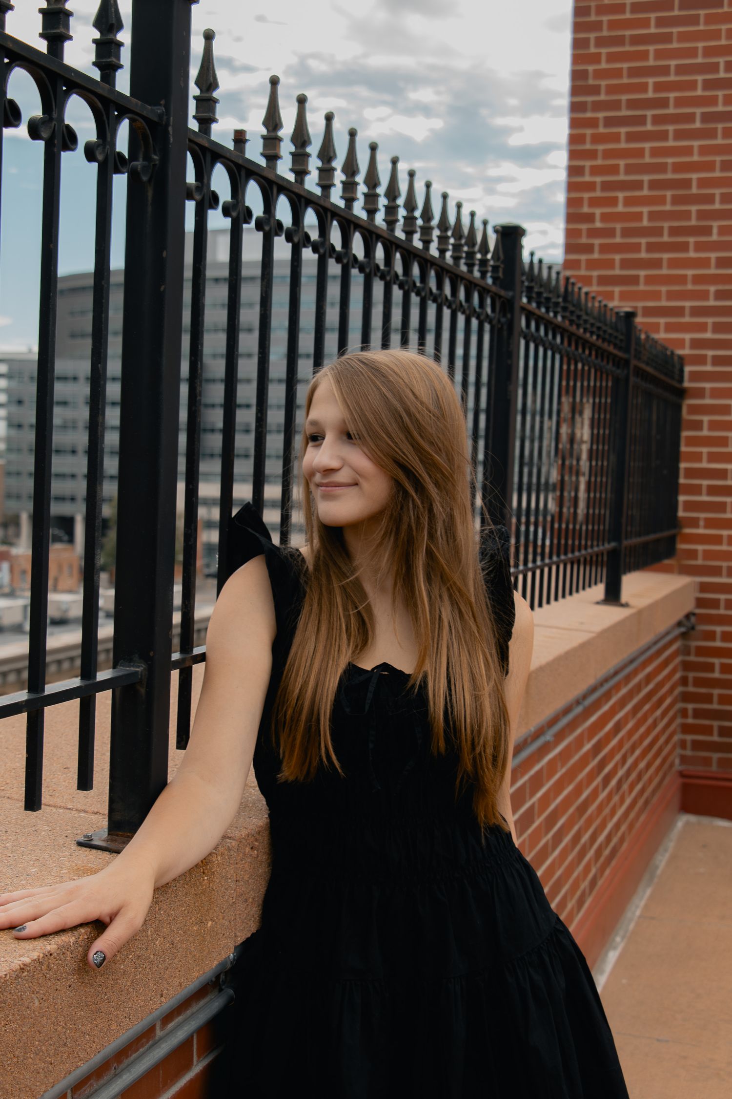 A woman in a black dress leans against an ornate iron railing on a brick terrace with cloudy skies in the background.
