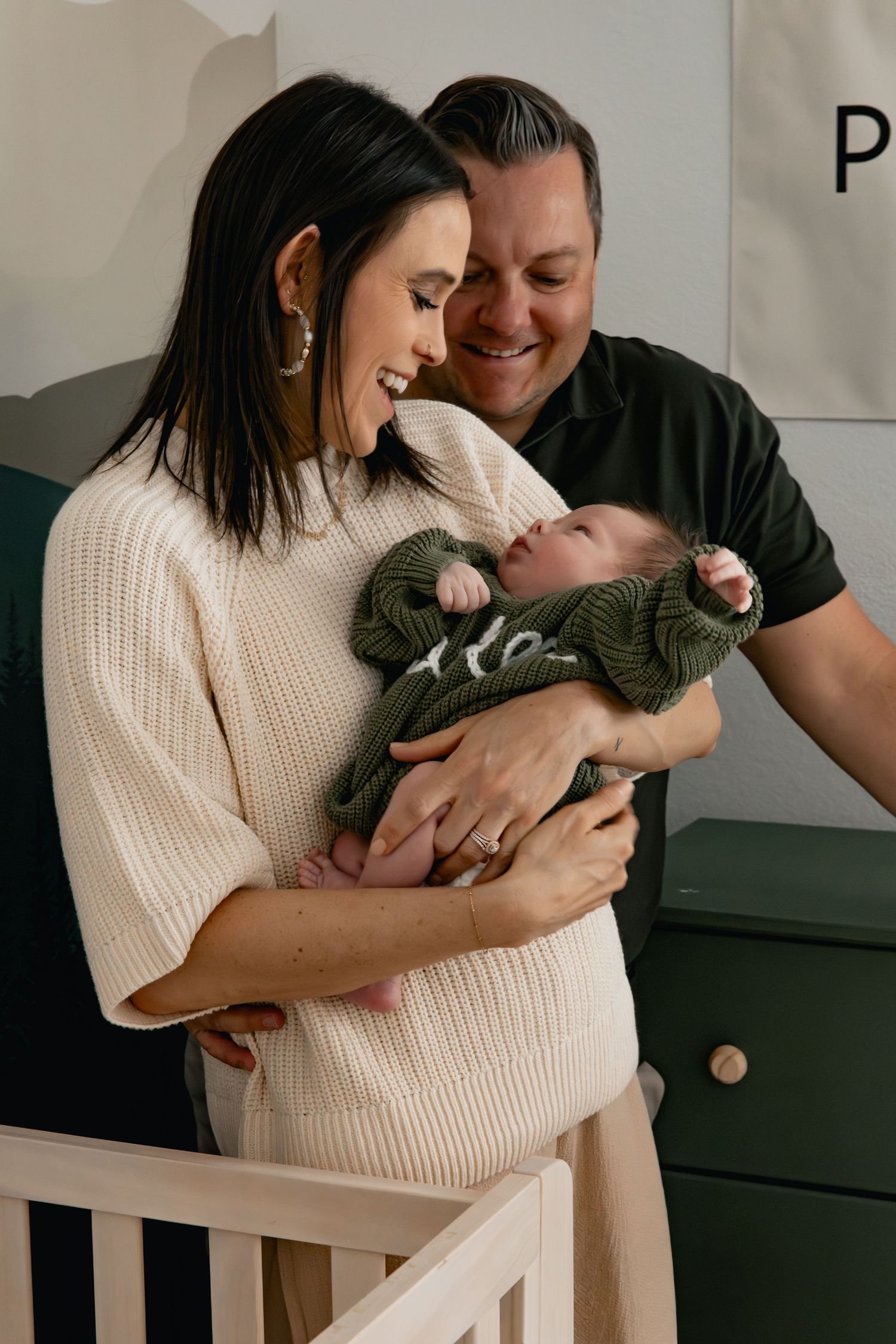 Happy parents smiling at their newborn baby dressed in green, standing by a white crib in a nursery.