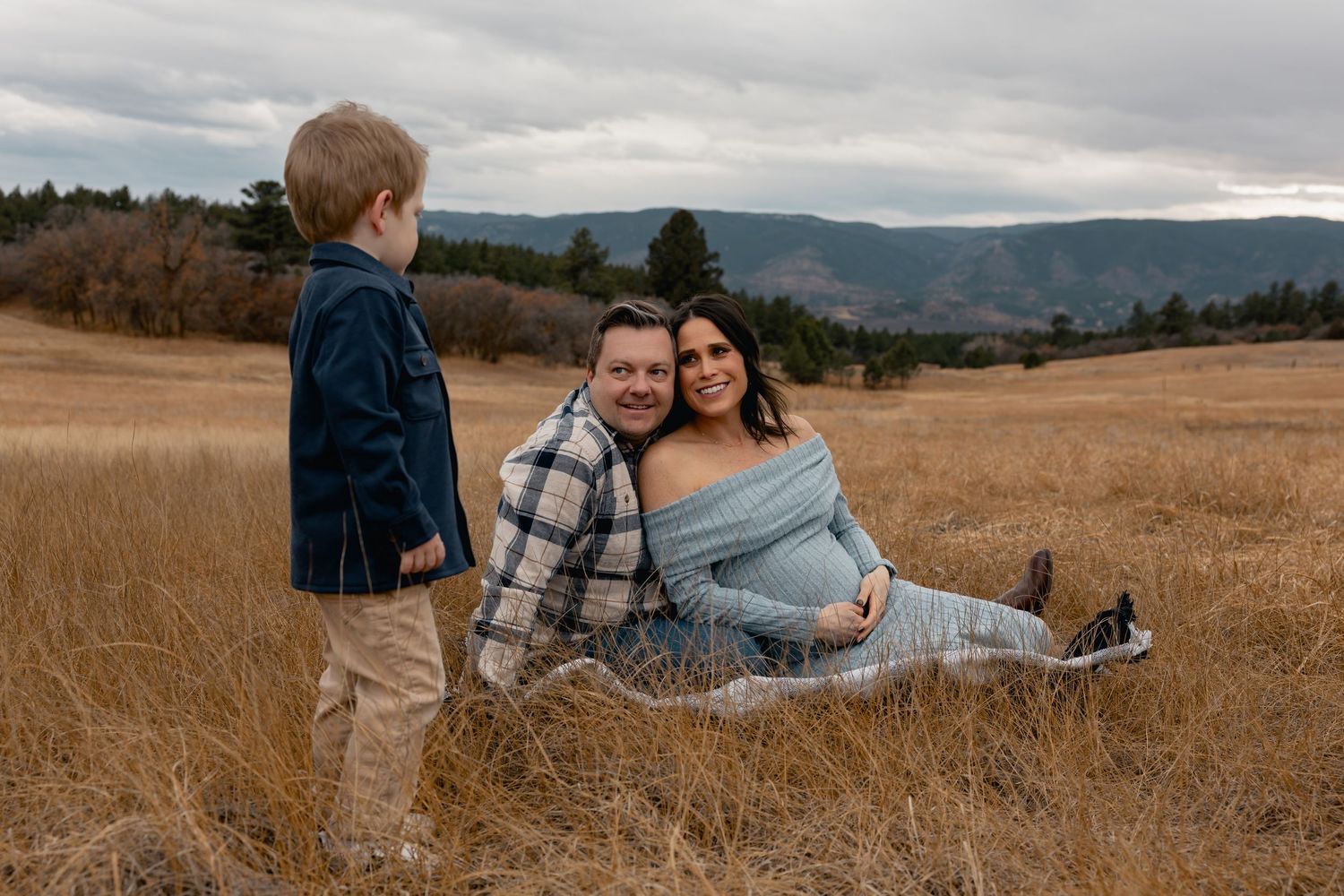 Pregnant woman and husband sit in golden field with toddler son, mountains in background, maternity photo.