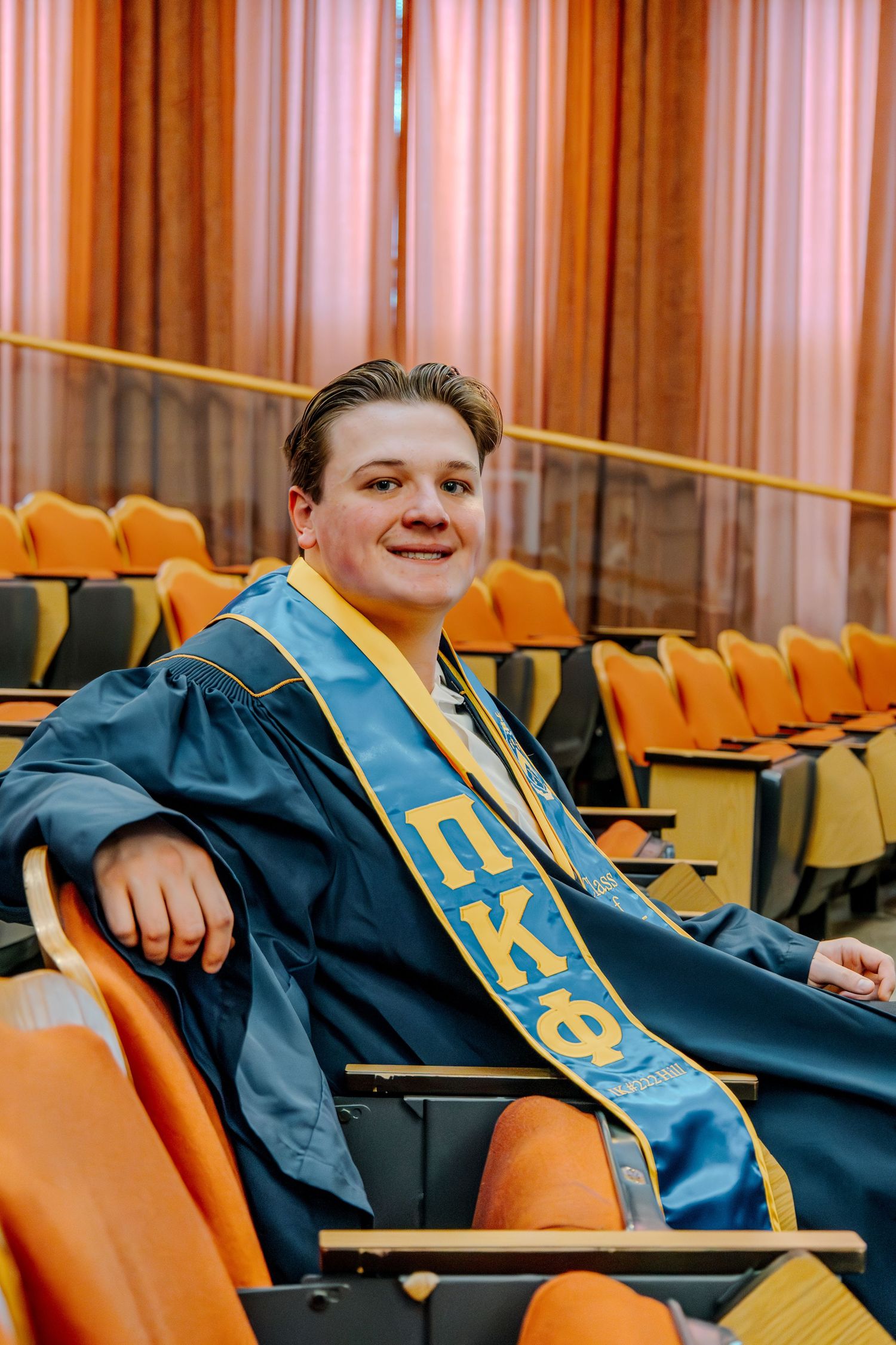 A graduate wearing a blue commencement gown and Pi Kappa Theta sash sits smiling in an auditorium with orange seats.