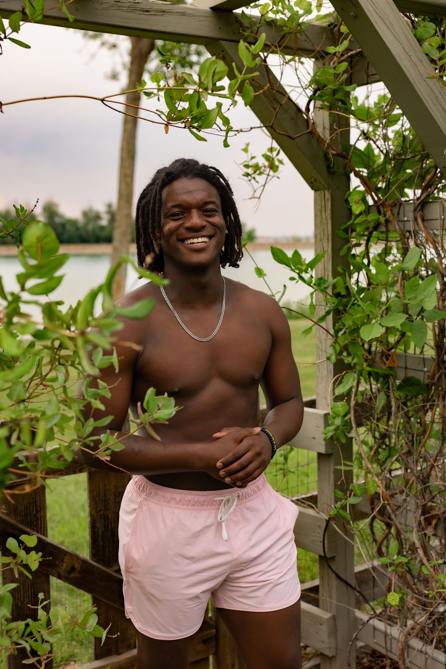 Smiling shirtless Black man with dreadlocks in pink shorts standing under a wooden garden arch with green vines.