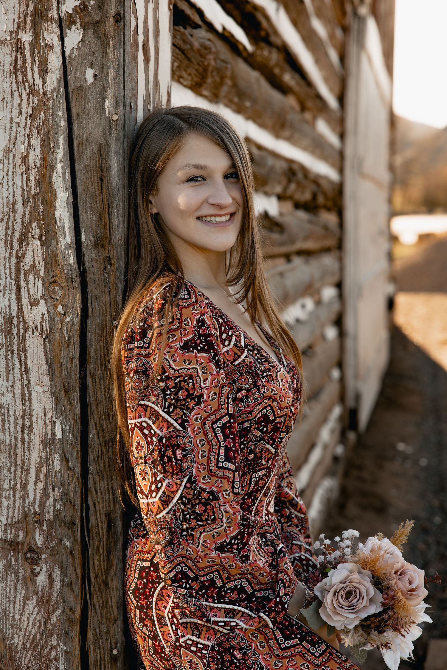 Young woman in boho paisley dress holding dried floral bouquet, leaning against rustic wooden barn wall, smiling outdoors.