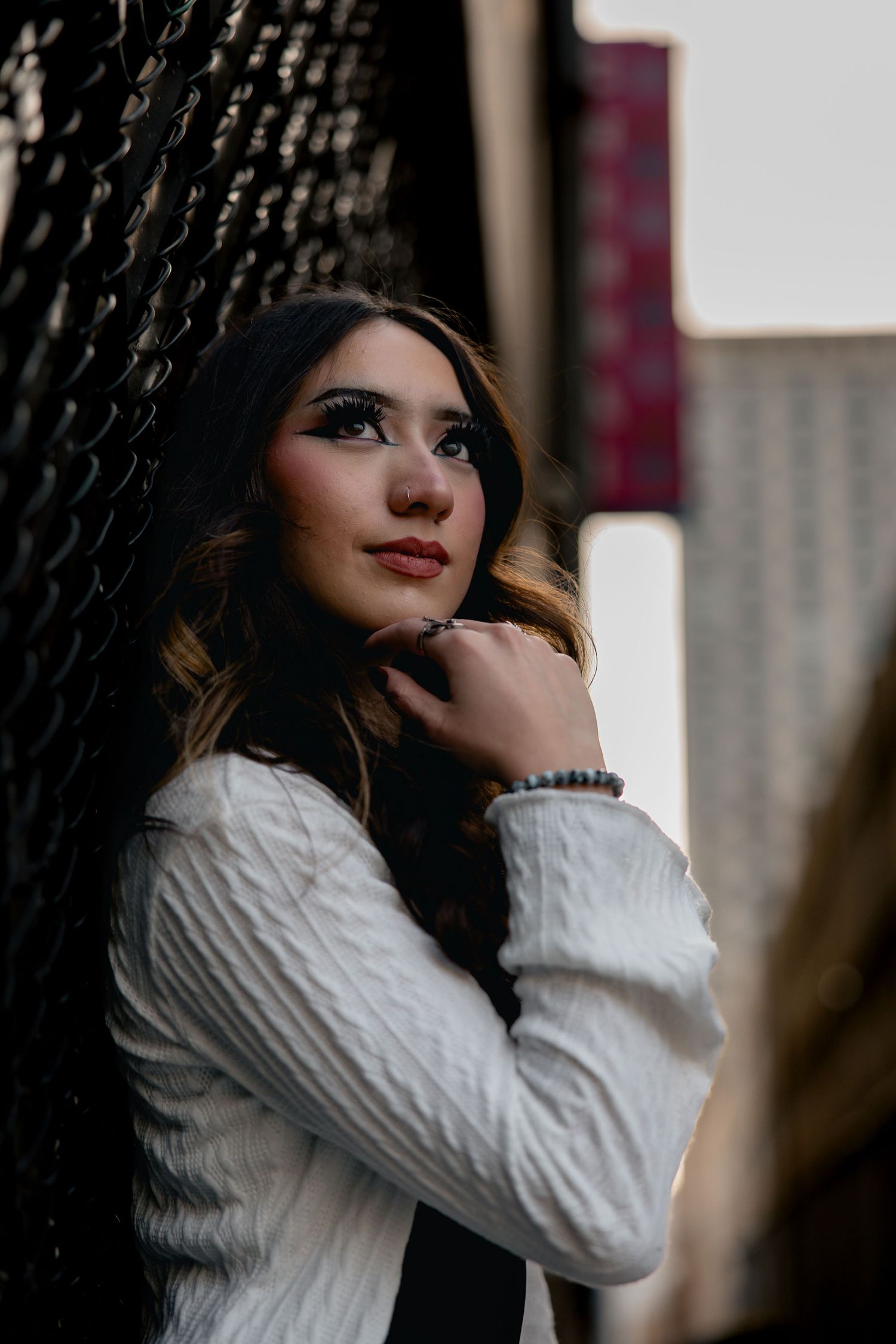 Young woman in white cable-knit sweater poses against chain-link fence in urban setting with dramatic makeup.