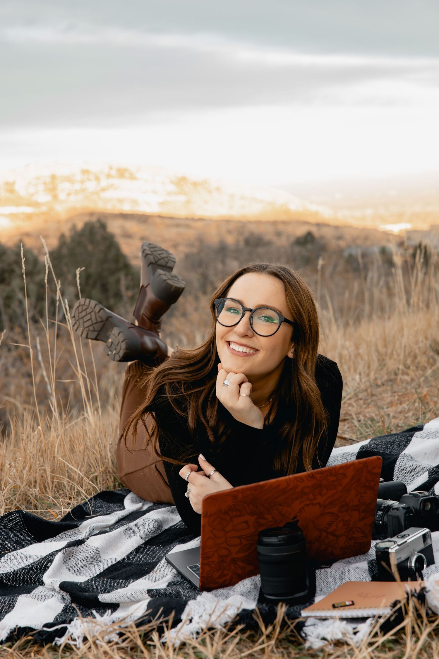 Person laying in autumn field with laptop wearing black sweater and glasses against scenic mountain backdrop.