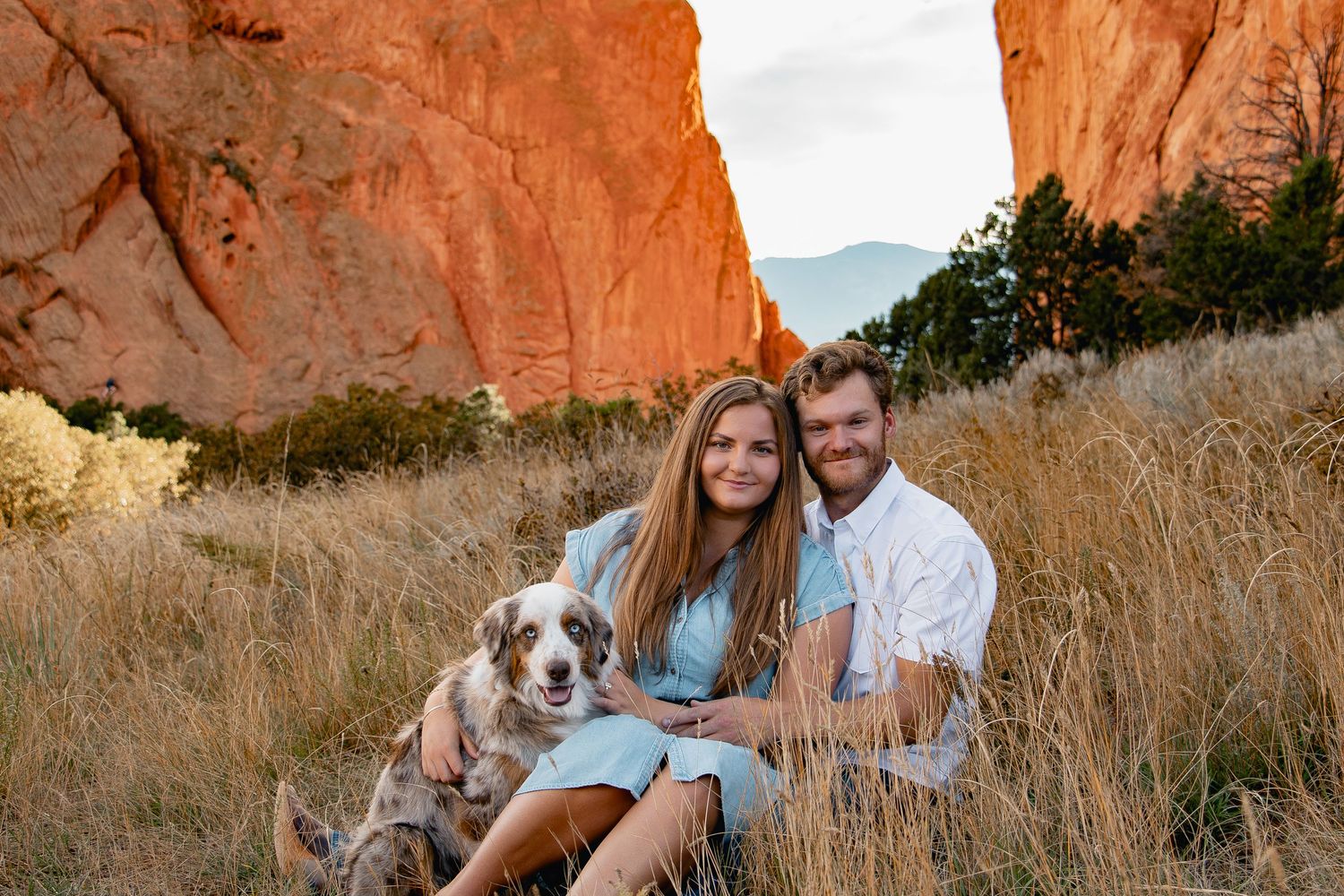 A couple sits together in a field with their dog against red rock formations during golden hour.