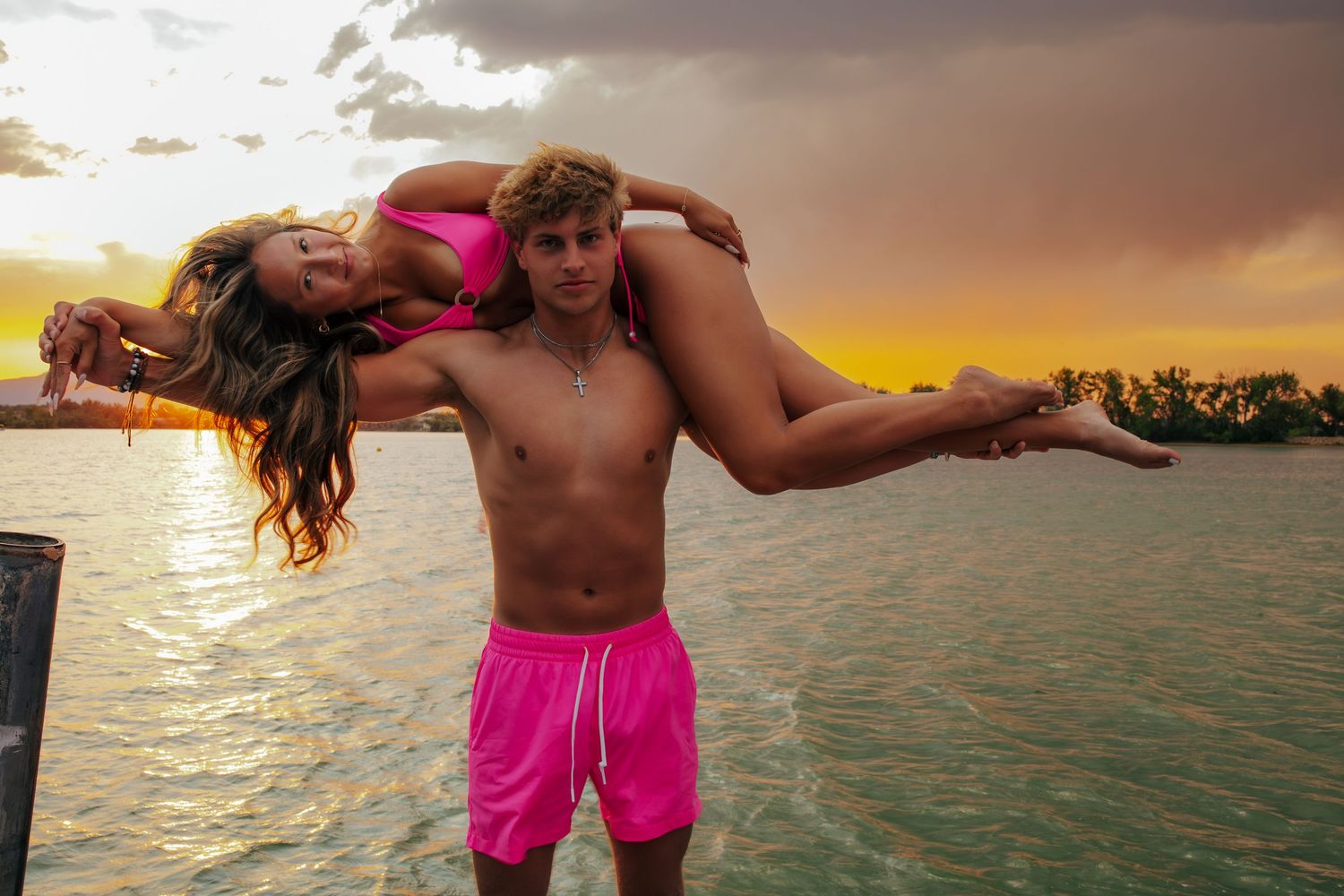 Couple in pink swimwear posing at sunset over tropical waters, man carrying woman on his shoulders.