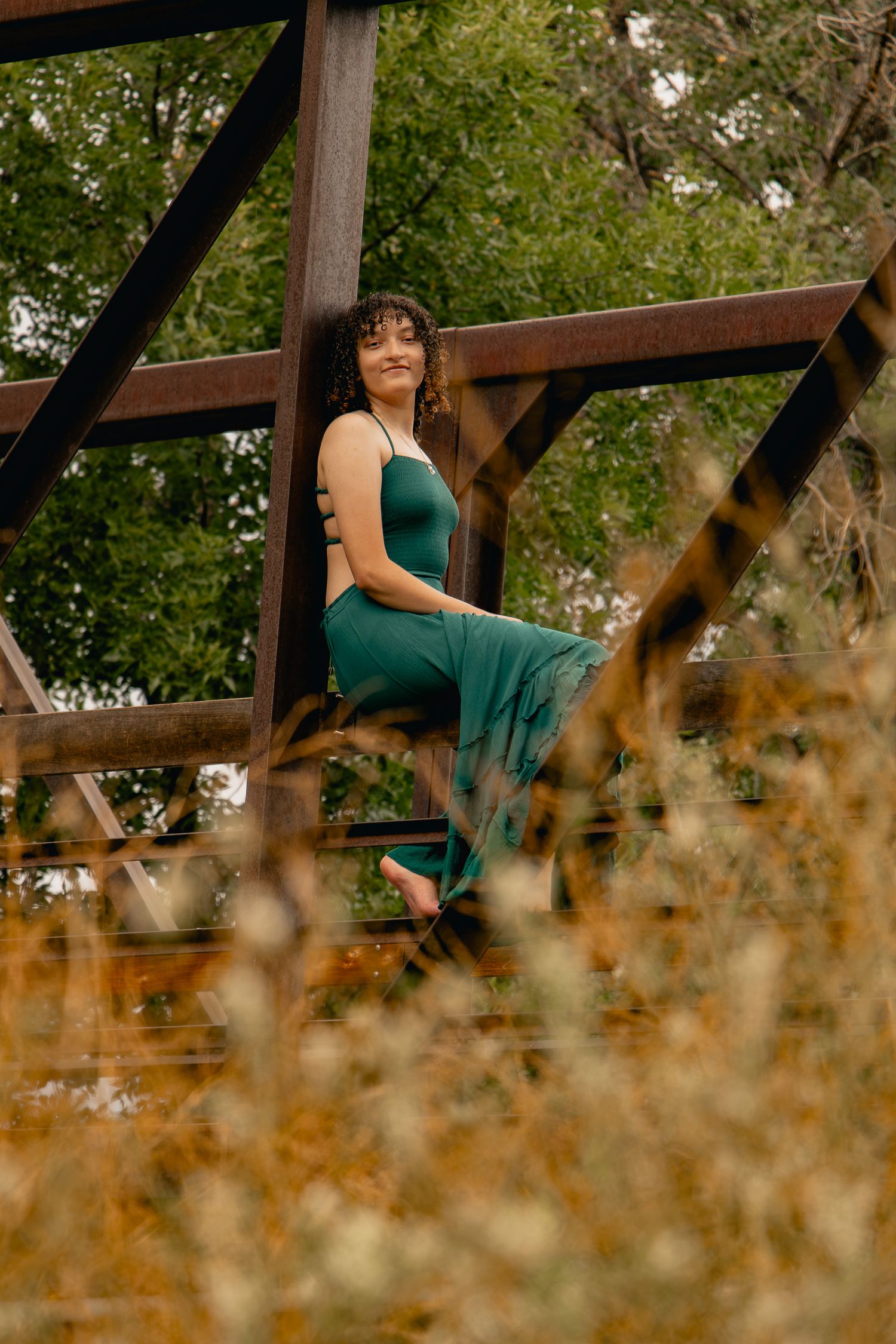 A person in a teal dress sits on a wooden deck railing surrounded by autumn foliage and warm sunlight.