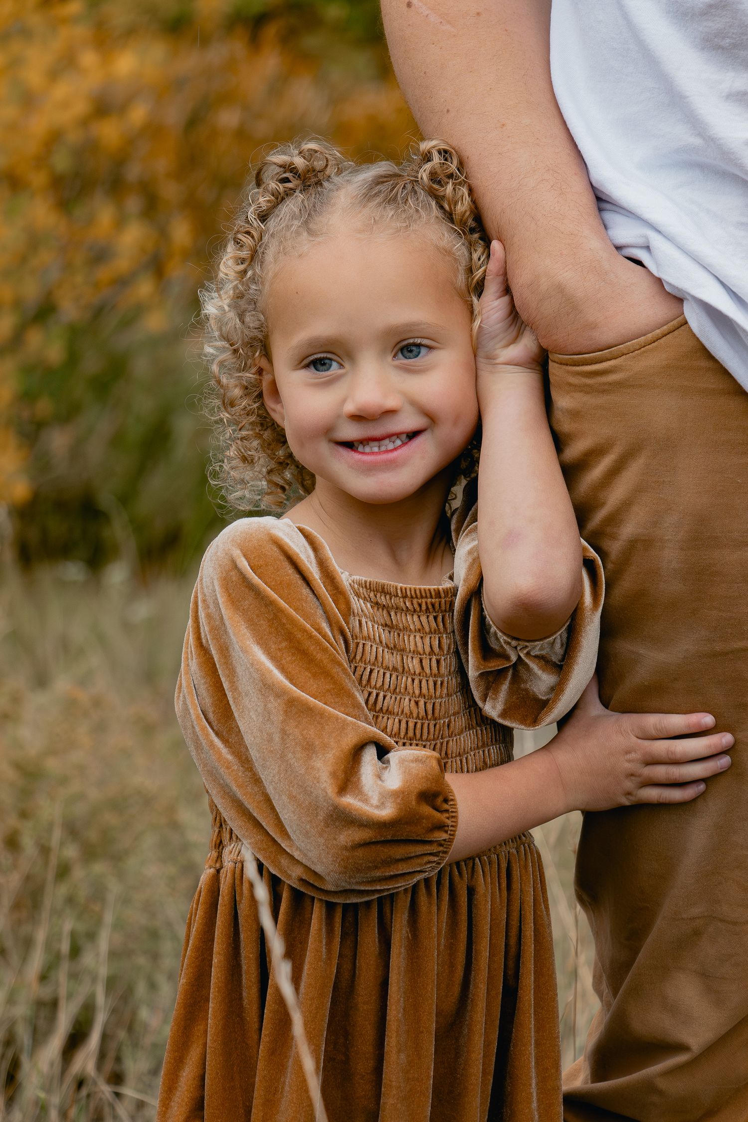Child in brown velvet dress with curly hair hugging parent's leg outdoors during autumn.