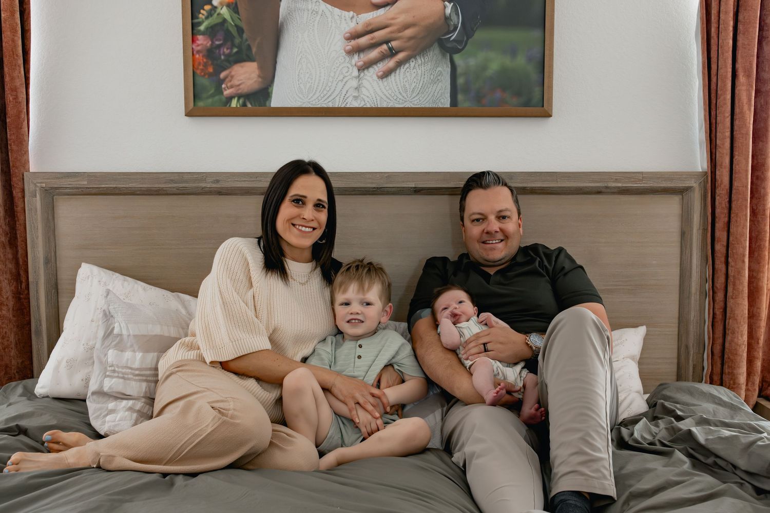 Happy family of four sitting on a bed, with parents holding two young children, beneath a framed maternity photo.