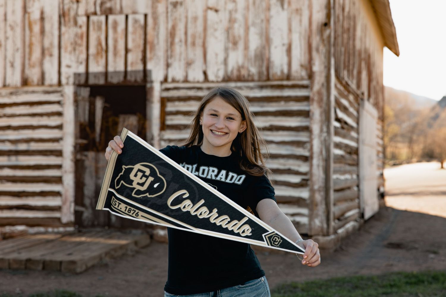 Smiling girl holding University of Colorado Buffaloes pennants wearing a CU black t-shirt outdoors near a barn.