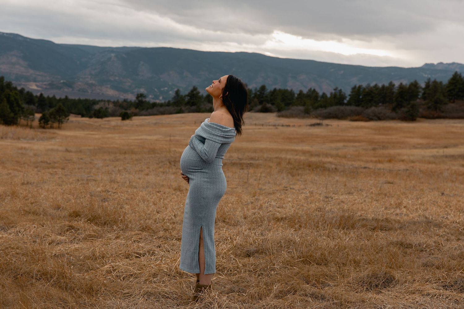 Pregnant woman in gray dress standing in golden field with mountains, head tilted back, cradling baby bump.