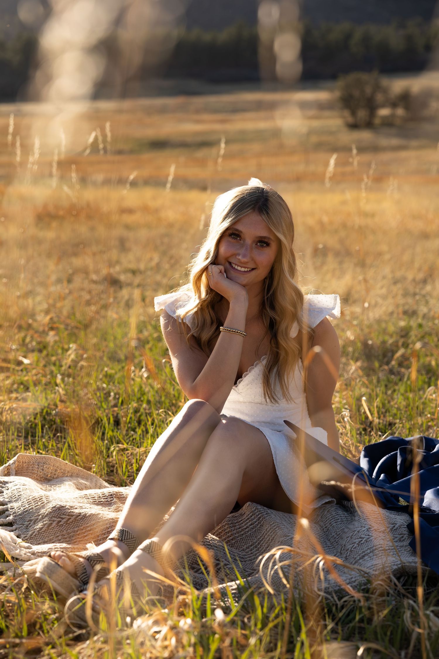 Smiling blonde woman in white dress sitting on blanket in golden sunlit meadow field during sunset.