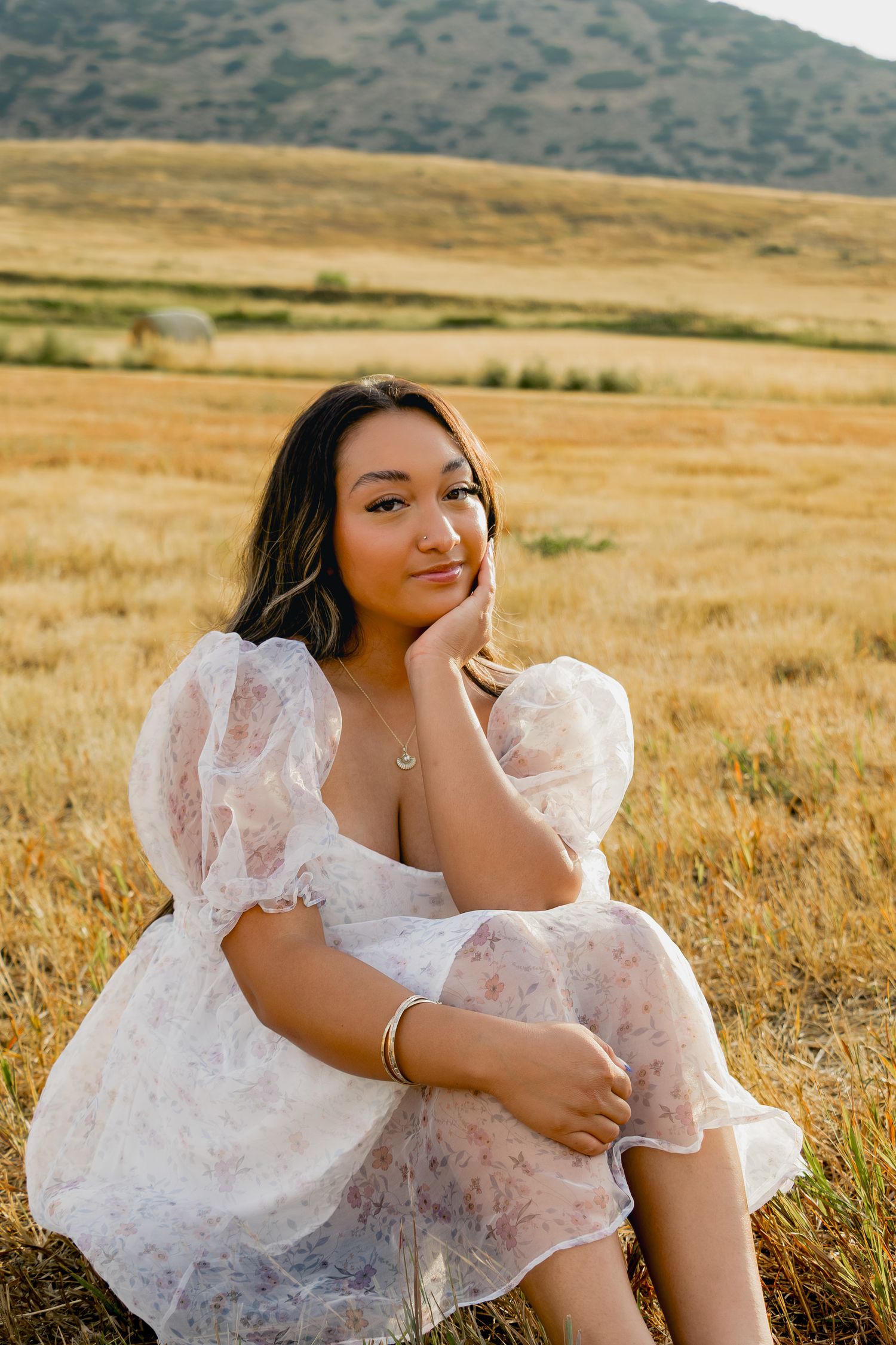 A person in a white lace dress sits gracefully in a golden field with mountains in the background.