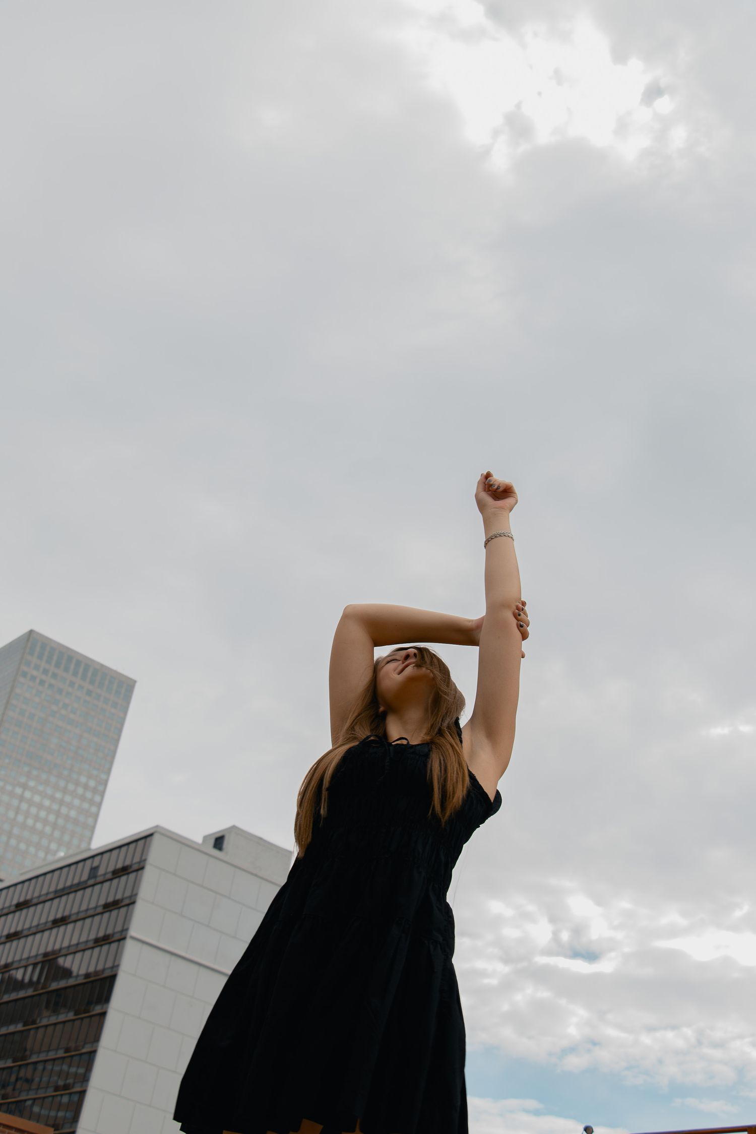 A figure in a black dress stretches upward against a cloudy urban skyline with tall buildings in the background.