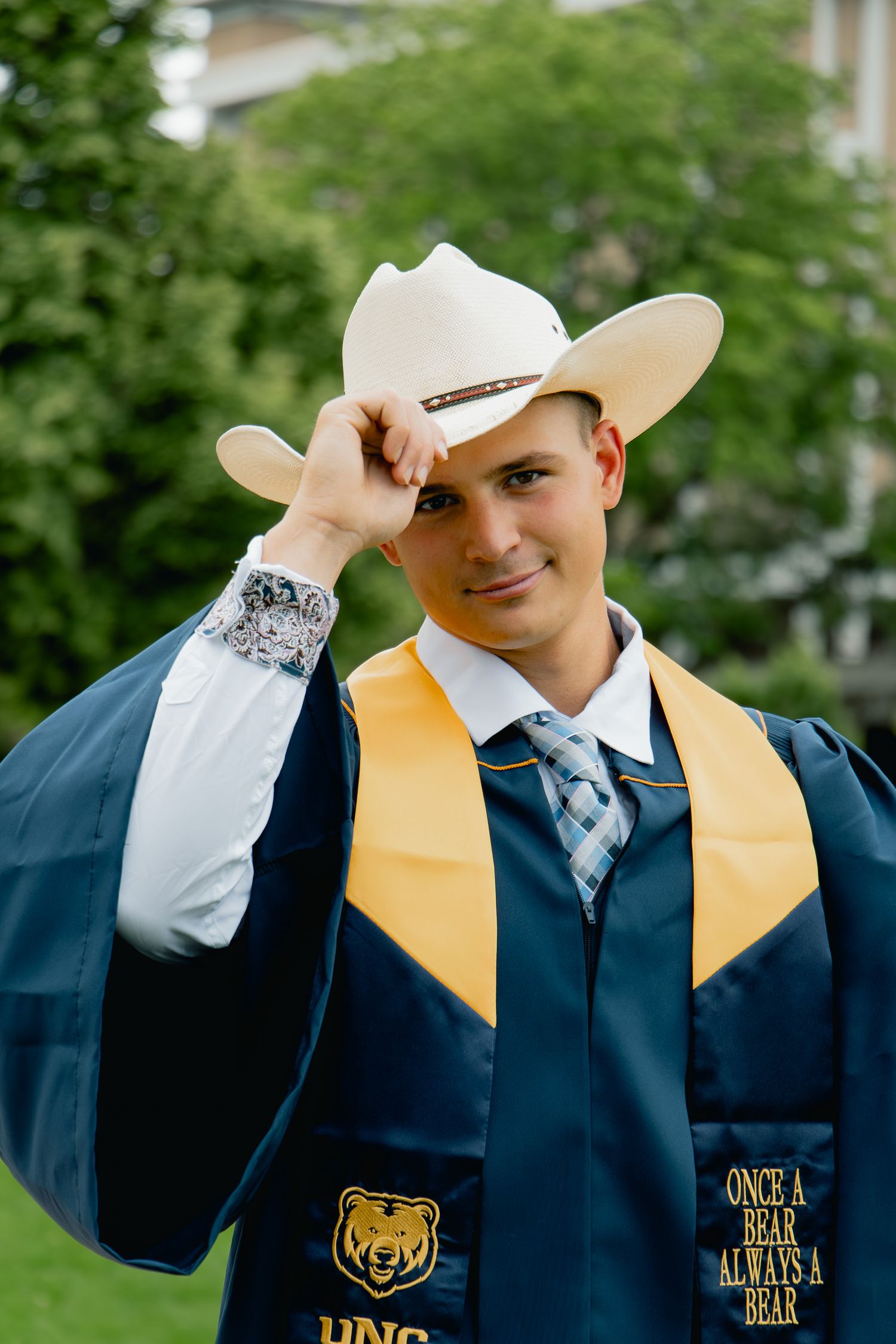 Graduate in navy blue gown with gold stole adjusts white cowboy hat in photo series.