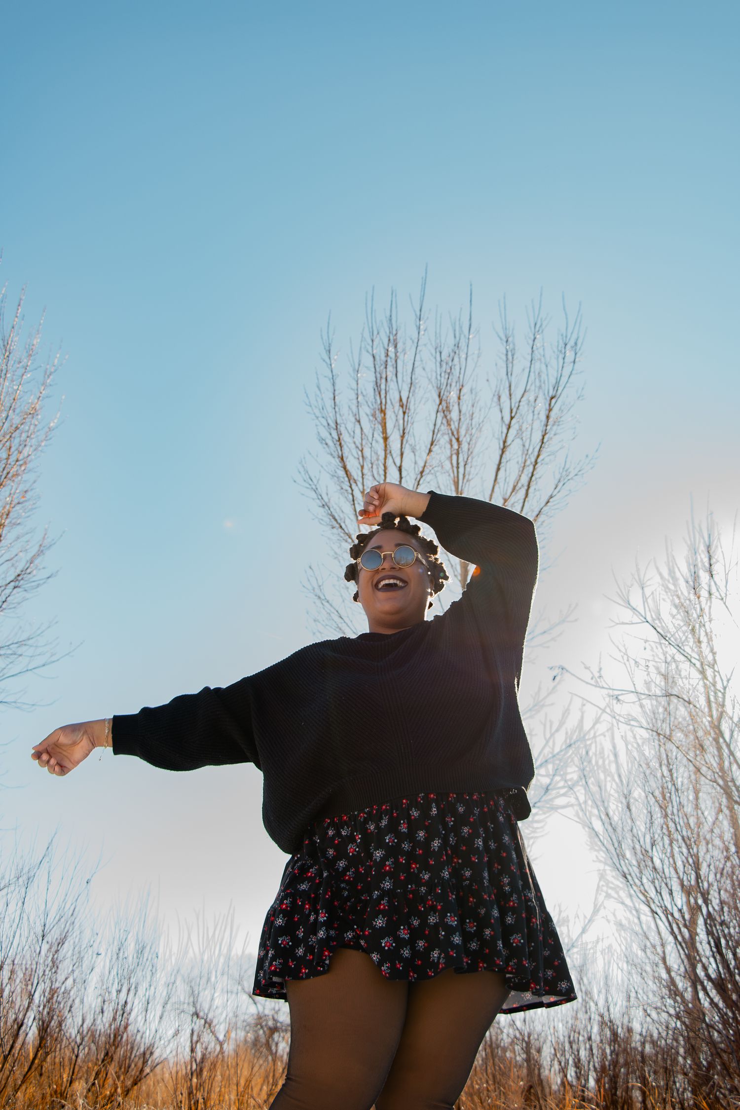 A joyful woman in a black sweater and floral skirt dancing outdoors against a blue sky with bare trees.
