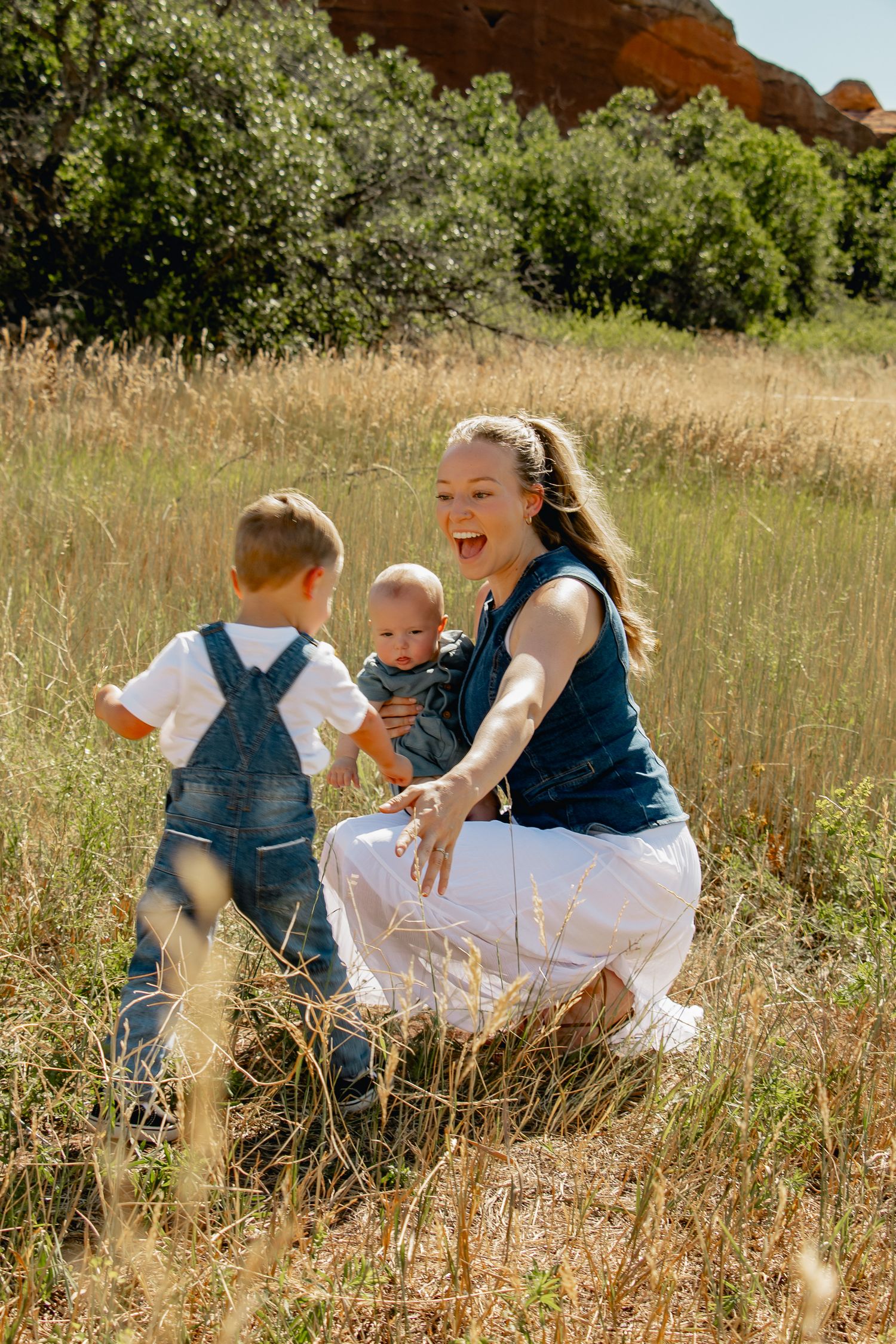 A family shares a joyful moment together in a sunlit field with tall grass and red rock formations in the background.