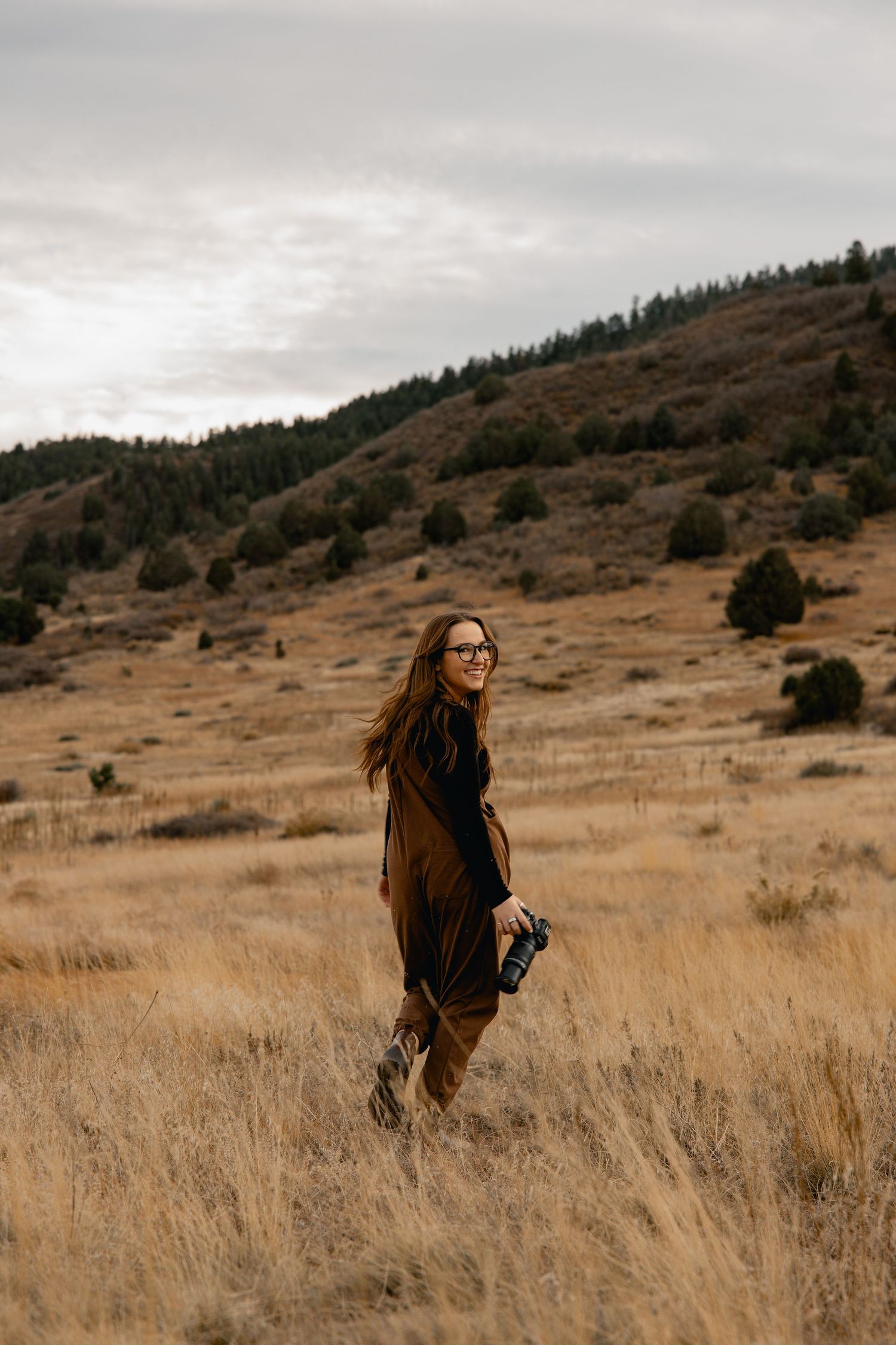 A person walks through tall golden grass on a hillside with evergreen trees and cloudy skies in the background.