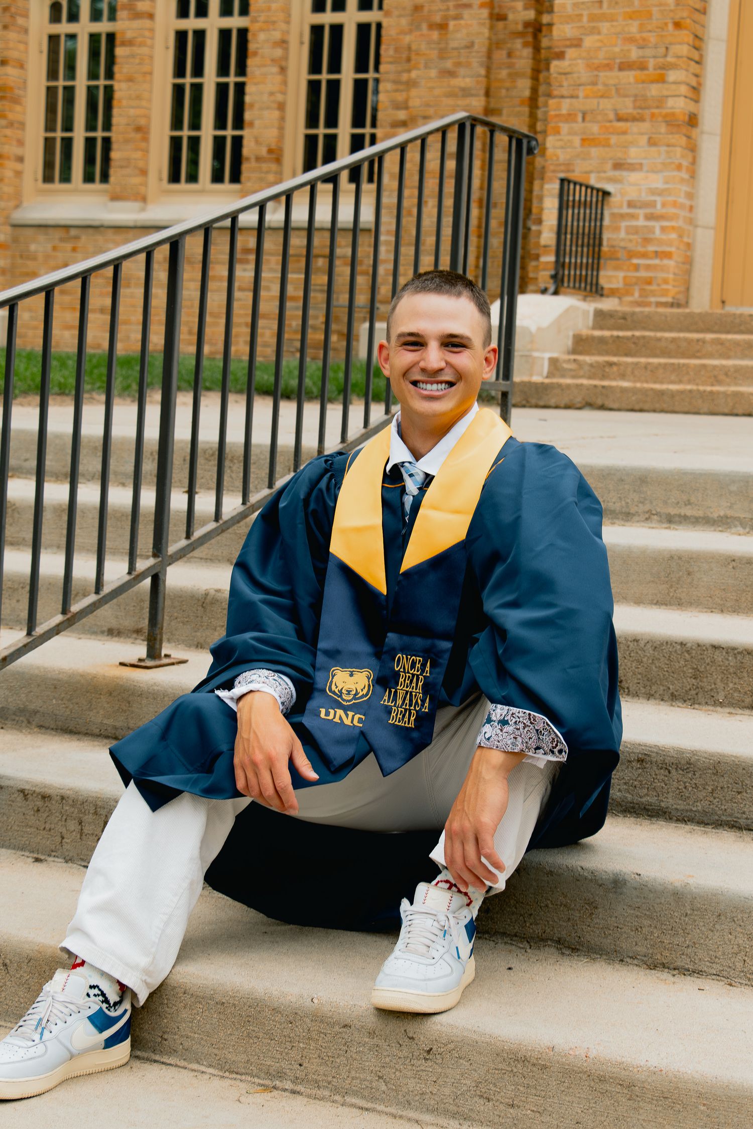 Someone wearing a blue graduation gown with gold stole and white sneakers sits casually on stone steps.