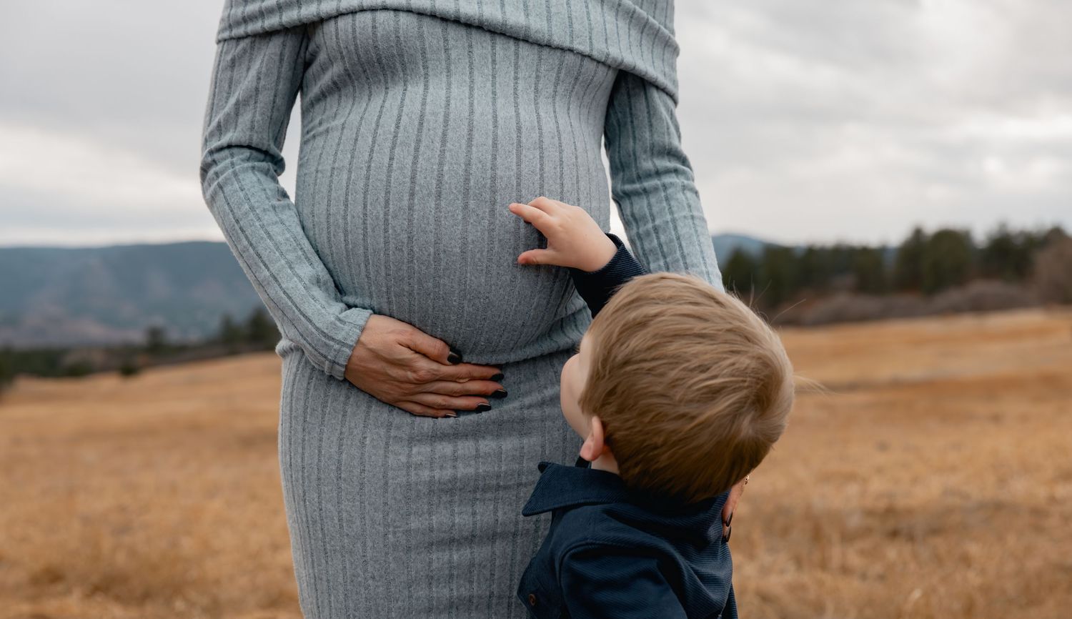 Young boy touching pregnant mother's belly outdoors in a golden field with mountains in the background.