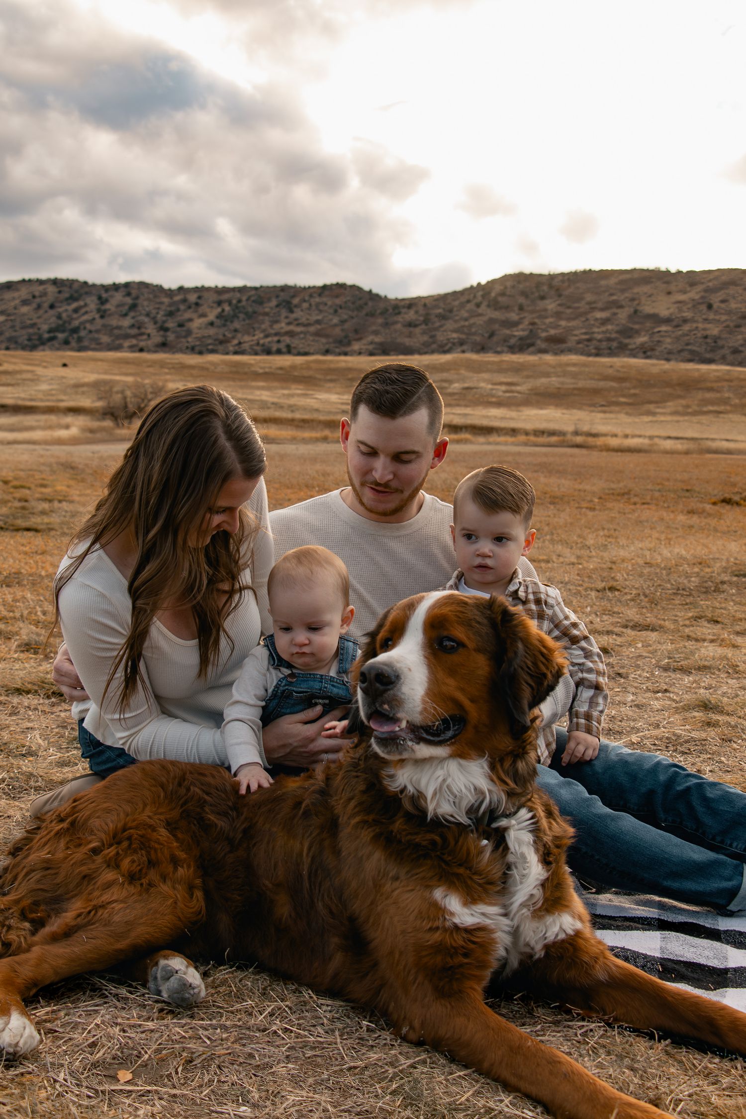 A family sits together on dry grassland with their large brown and white dog while mountains loom in the background.