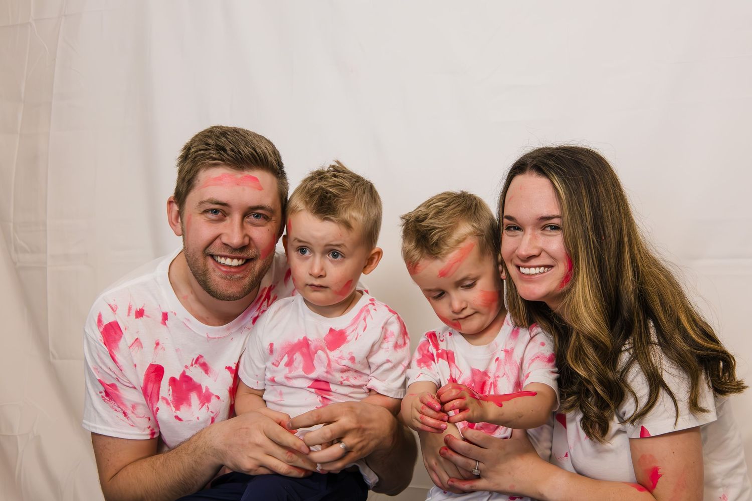 Family wearing matching white and pink tie-dye shirts sit on floor smiling while cuddling together.