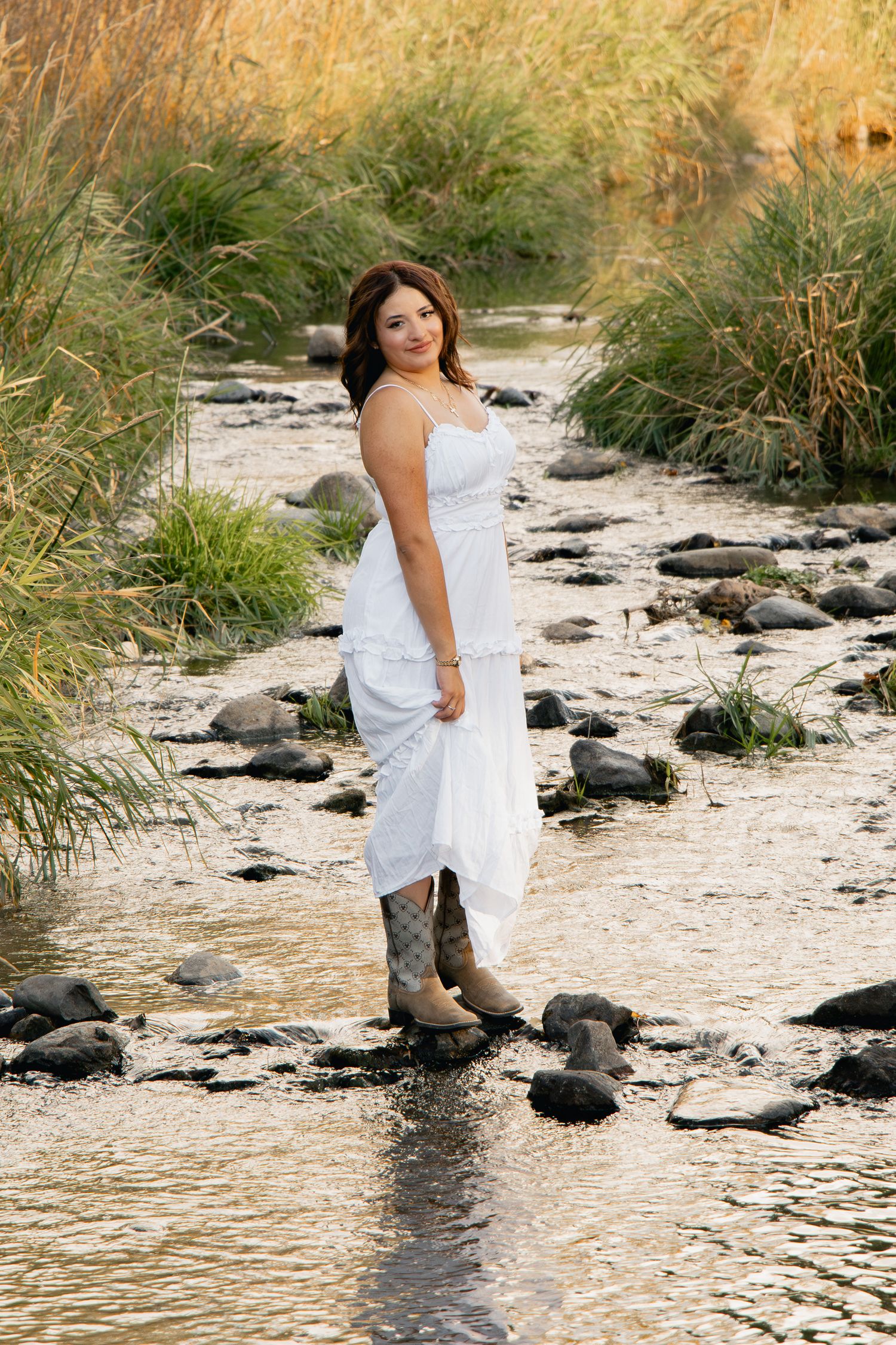 A person in a white dress sits by a rocky stream during golden hour with tall grass in the background.