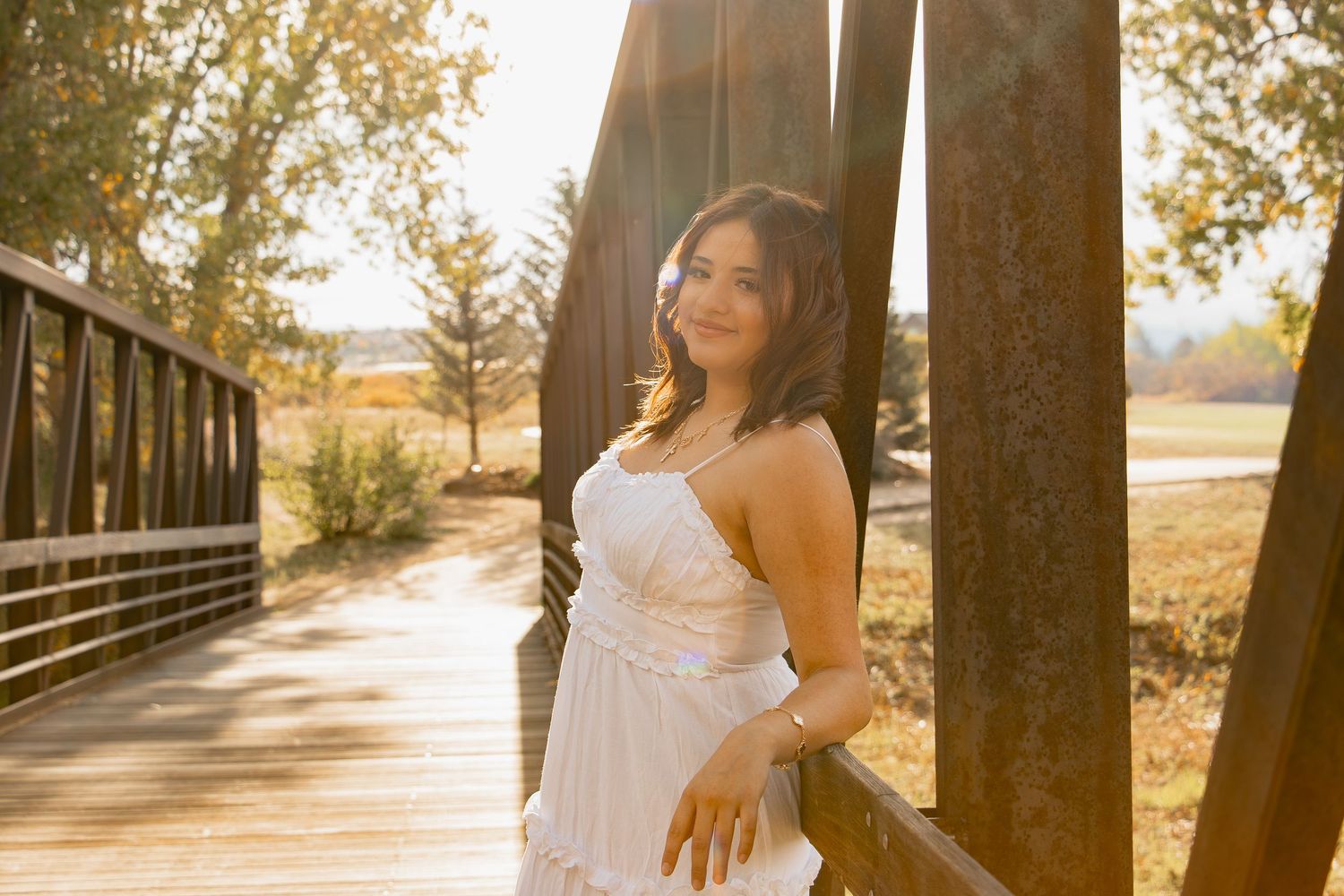 A woman in a white dress poses along a wooden bridge during golden hour sunset.