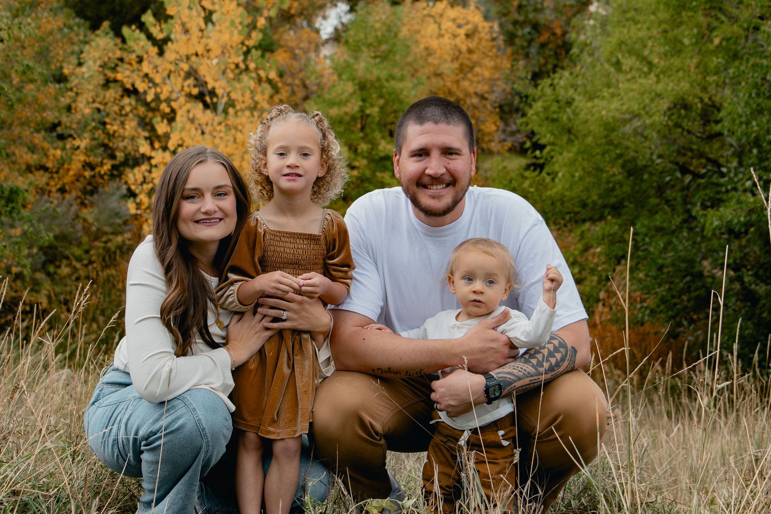 A family enjoys a warm autumn day together sitting in a golden field surrounded by trees.