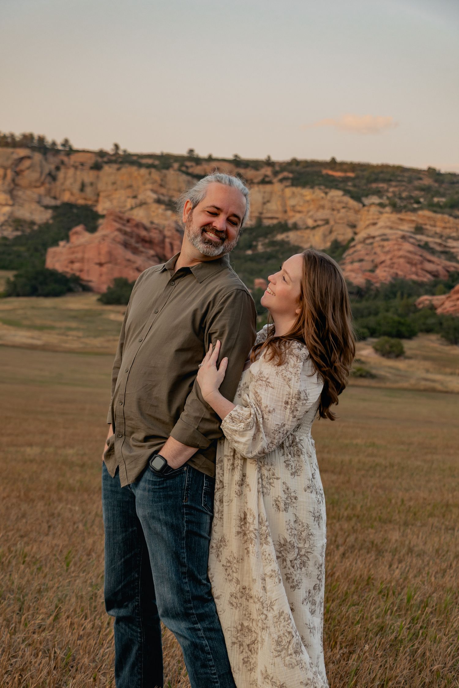 A couple laughs together in front of scenic red rock formations during golden hour in a grassy field.