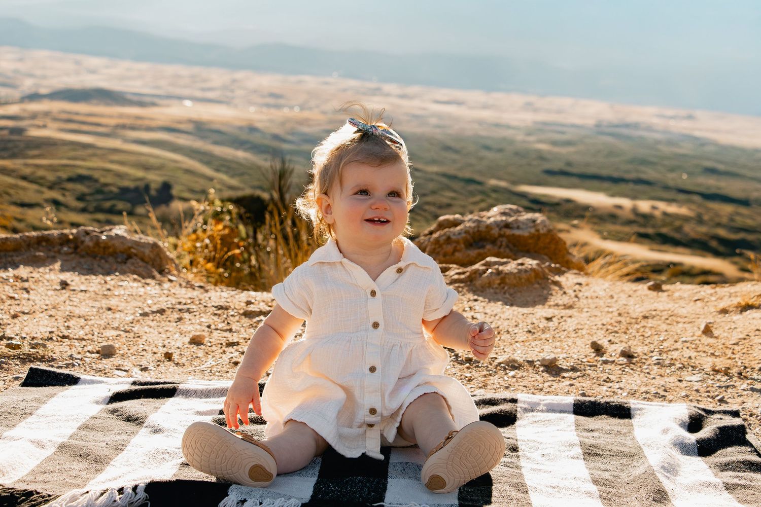 A baby in white clothing sits on a blanket outdoors during golden hour with scenic mountain views in the background.