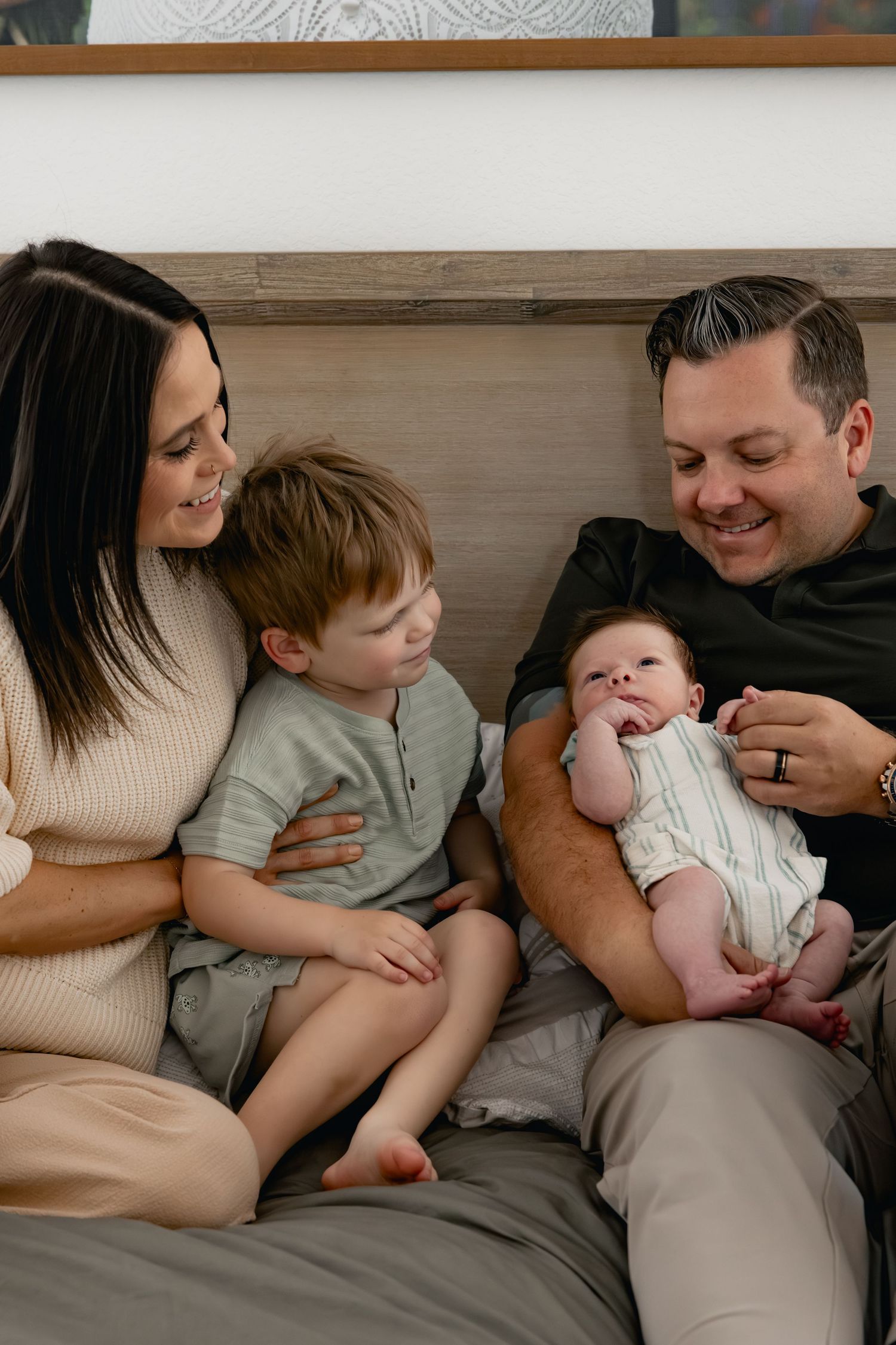 A happy family of four sits on a bed, parents smiling at their toddler and newborn baby in a warm home setting.