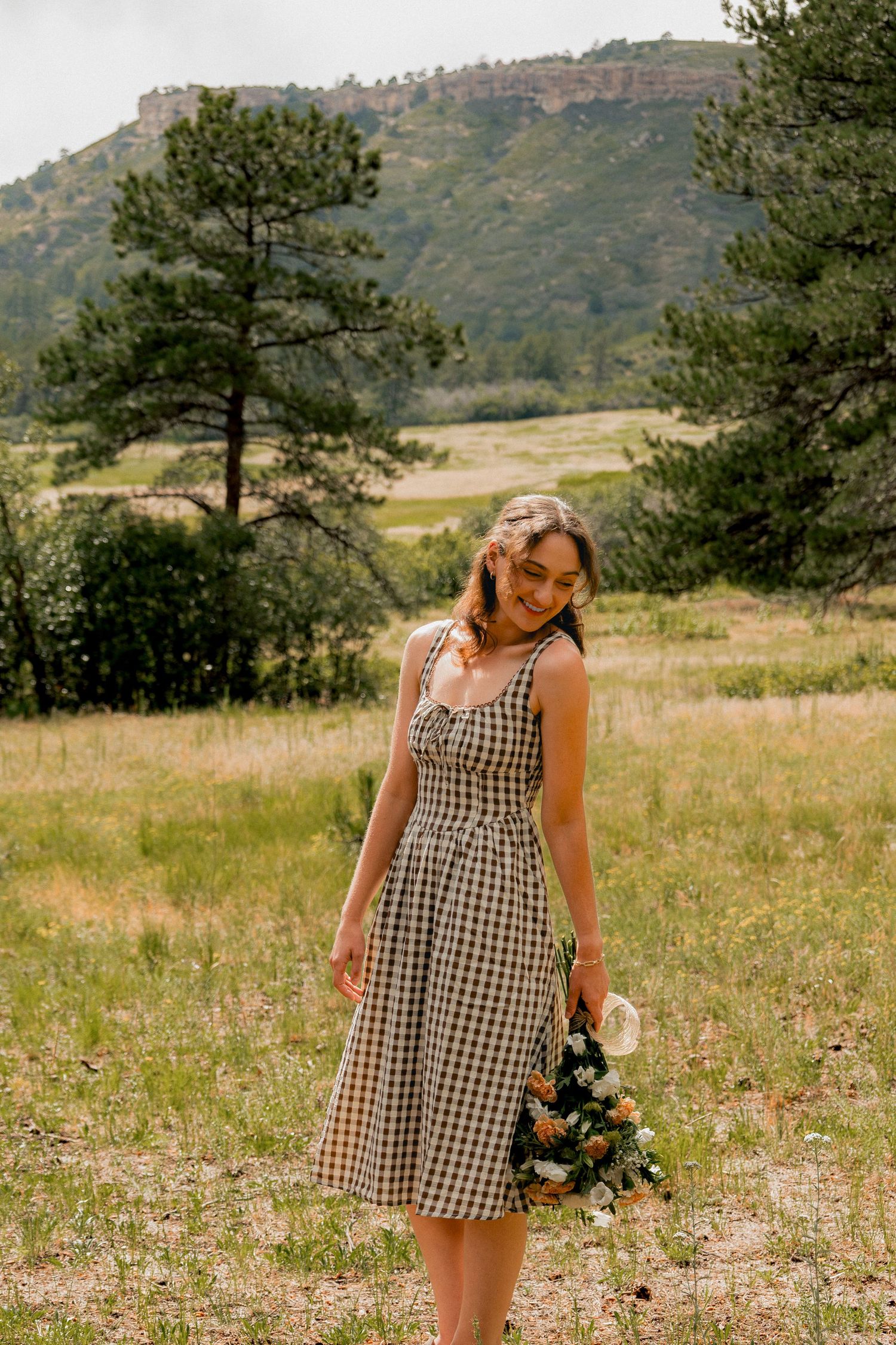 A person in a gingham dress stands outdoors against a scenic mountain landscape with evergreen trees.