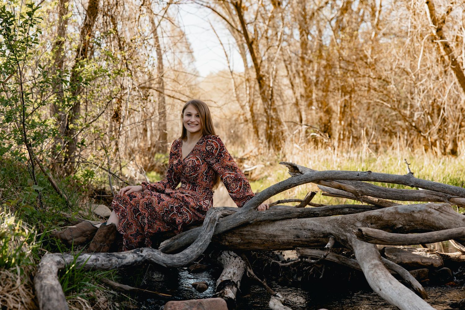 Woman in floral dress sitting on fallen logs in a sunlit forest setting.