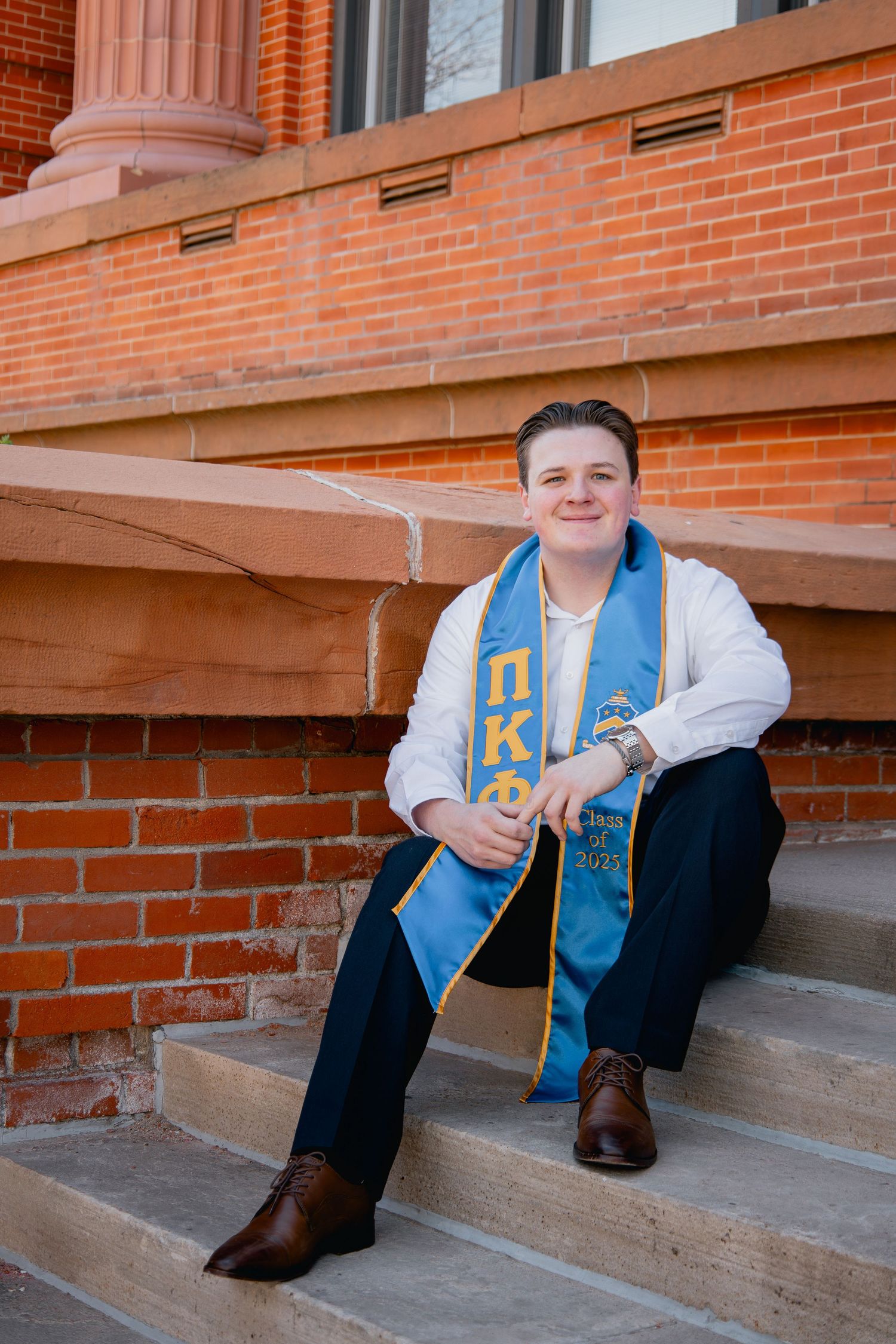 Graduate wearing a light blue Pi Beta Phi stole and white sweater sits on brick steps of a building.