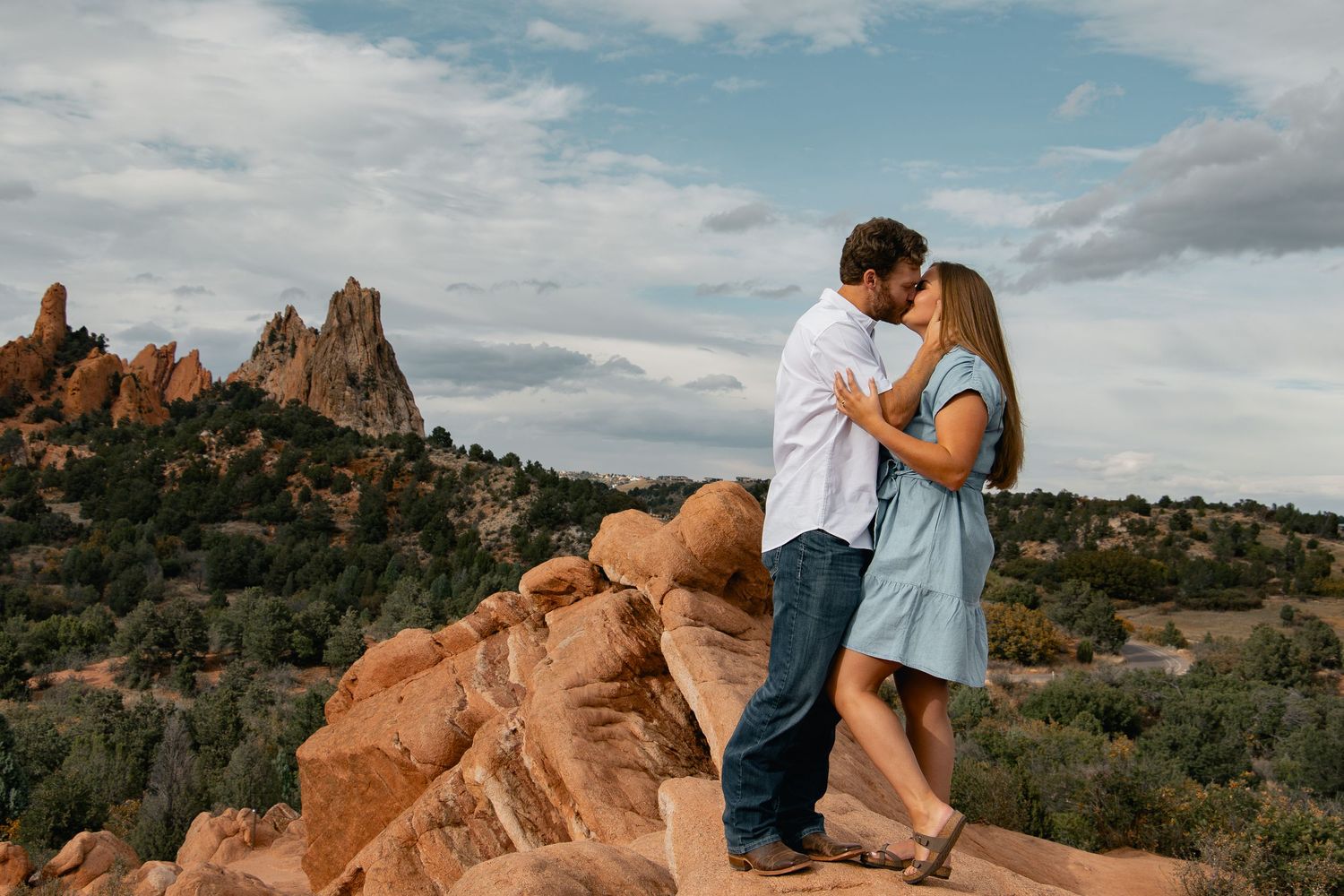 Two people embrace while sitting on red rock formations in a desert landscape with dramatic cloudy skies.