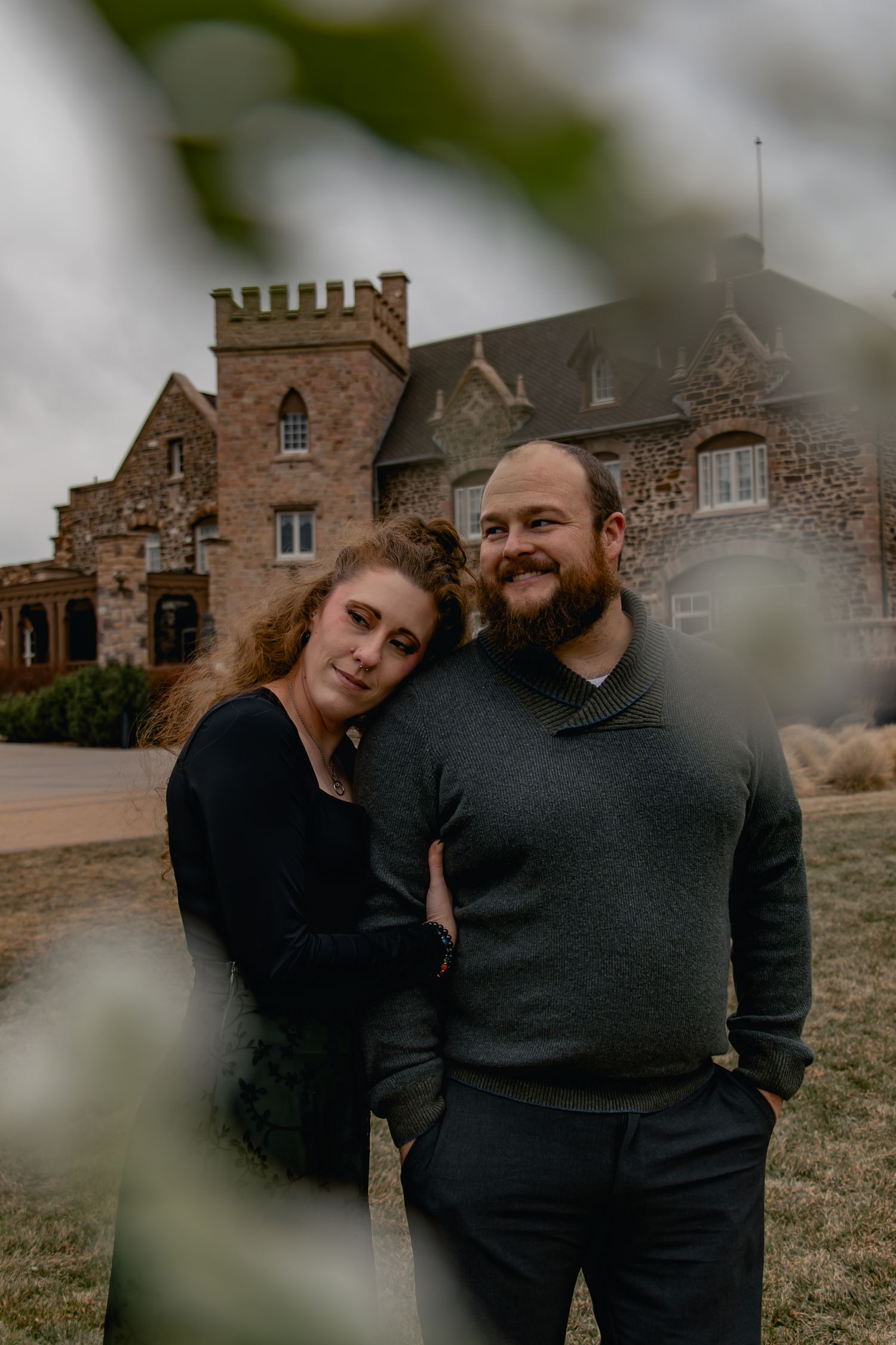 A couple shares intimate moments in front of a rustic stone building on an overcast day.