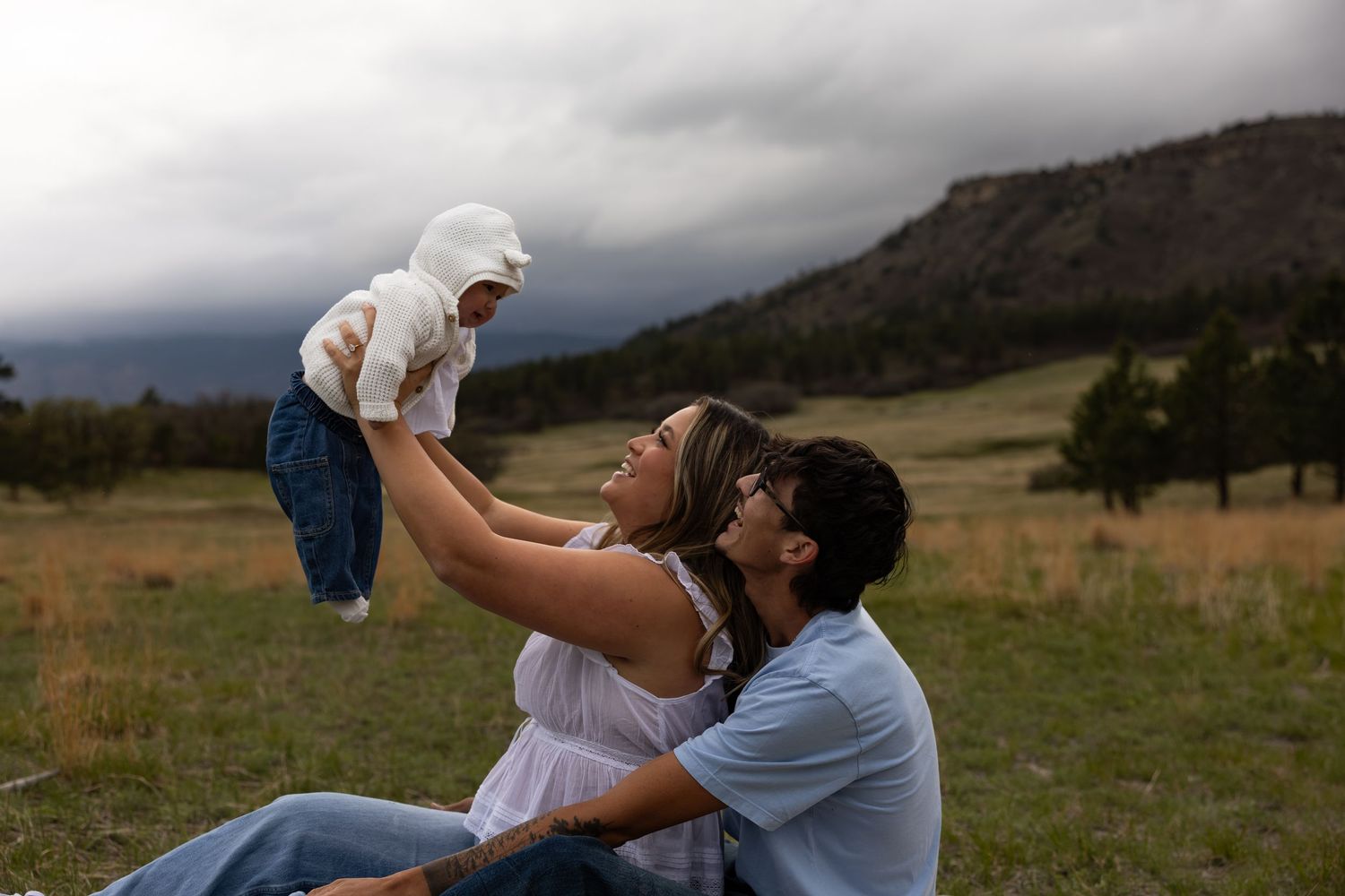 A young couple sits in a green meadow with mountains, lifting their baby in the air under a dramatic cloudy sky.