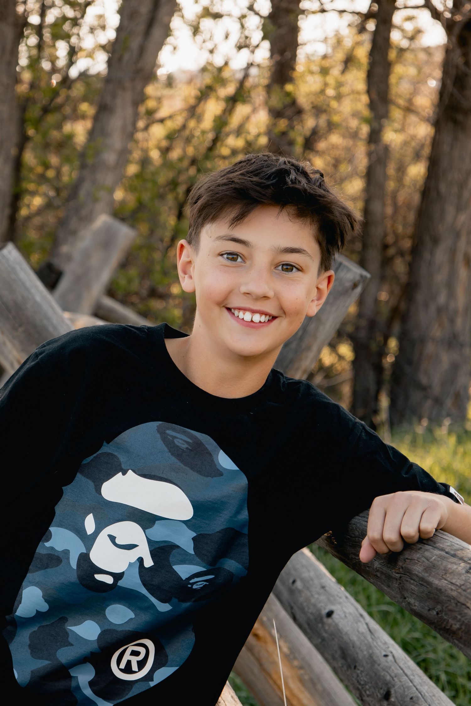 A smiling youth wearing a black sweatshirt with blue camo graphic design leans against wooden fence posts in forest setting.