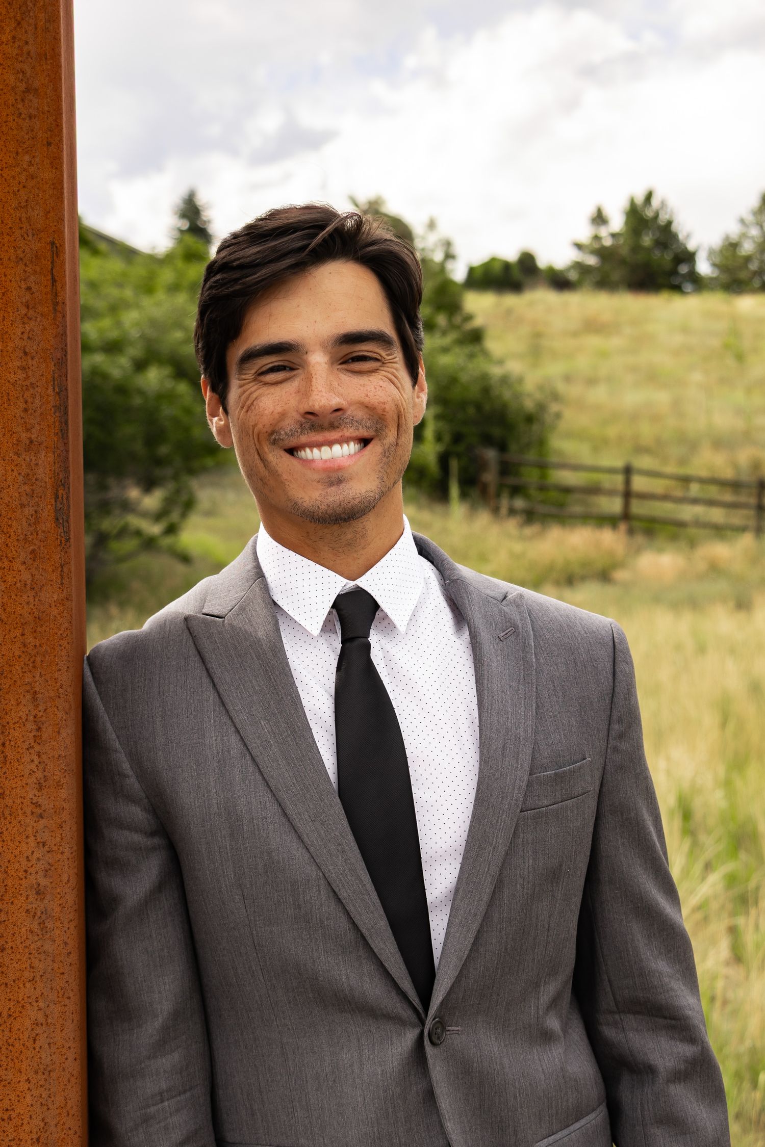 A well-dressed person in a grey suit and black tie smiling outdoors with a pastoral landscape in the background.